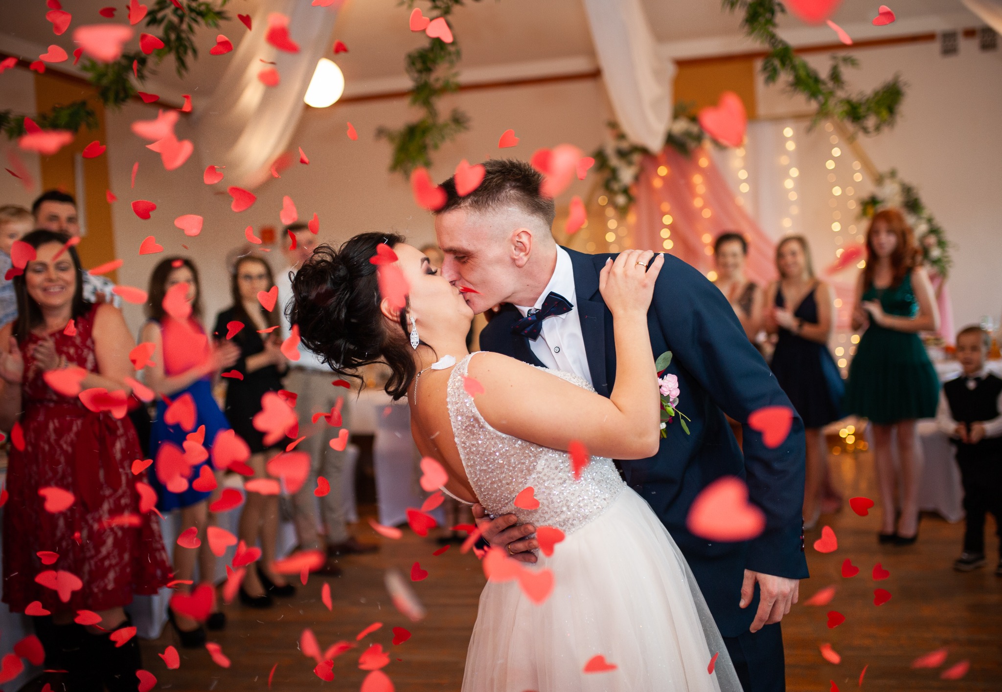 Pocałunek pary młodej w deszczu czerwonych serc podczas wesela. Goście klaszczą w tle, dekoracje świetlne. Ujęcie z bliska, ciepłe światło.