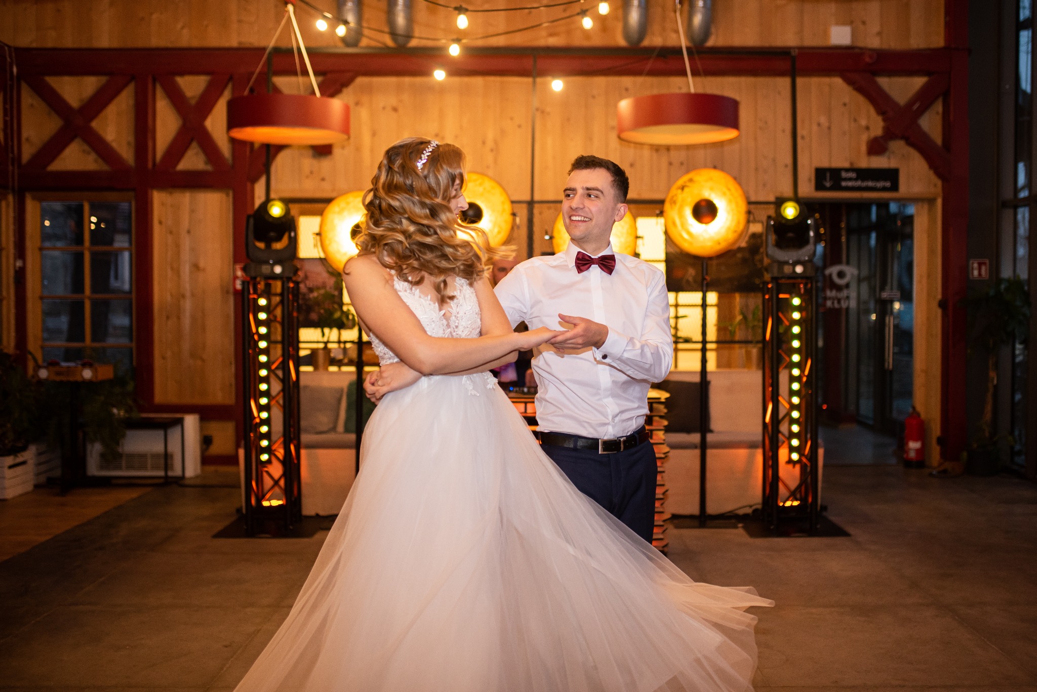Uśmiechnięta para młoda w tańcu, on w muszce, ona w sukni ślubnej z tiulowym dołem, na tle dekoracji sali z oświetleniem i drewnianymi elementami.