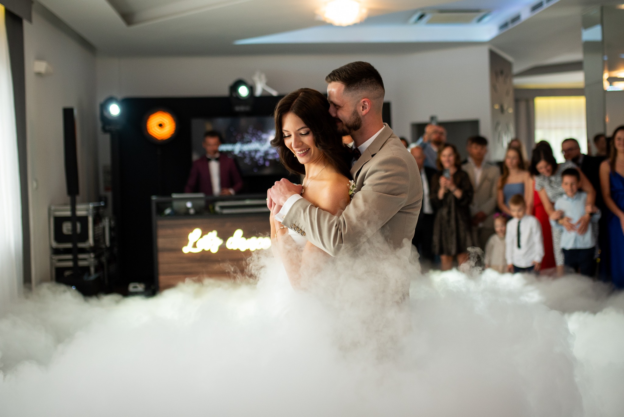 Uśmiechnięta para młoda w objęciach tańczy w obłokach dymu na tle gości i DJ-a z podświetlanym napisem 'Let's dance'.