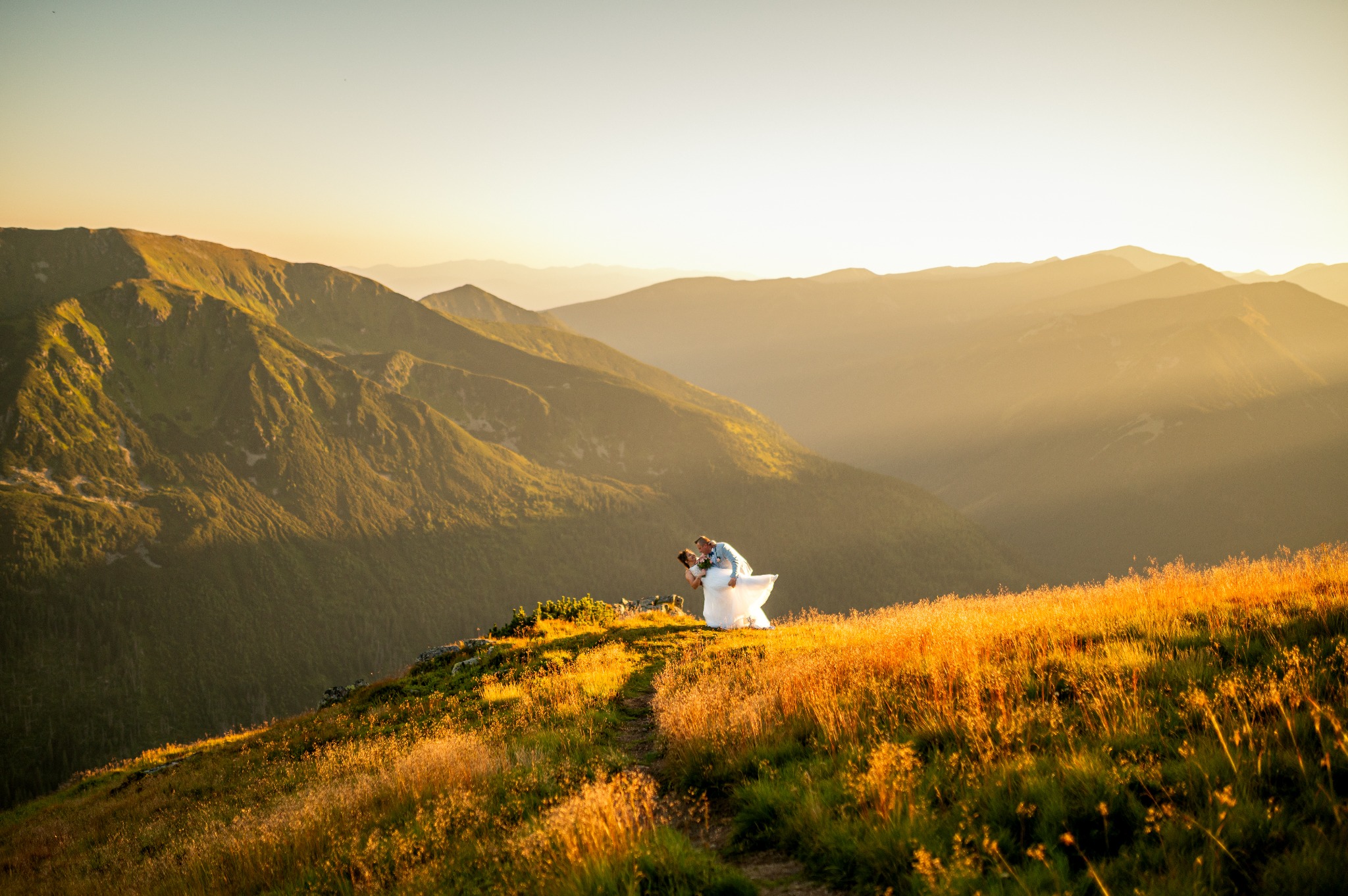 Para młoda w objęciach na tle gór o zachodzie słońca. Romantyczna sceneria, złote światło podkreśla uroczystość. Suknia ślubna powiewa na wietrze.