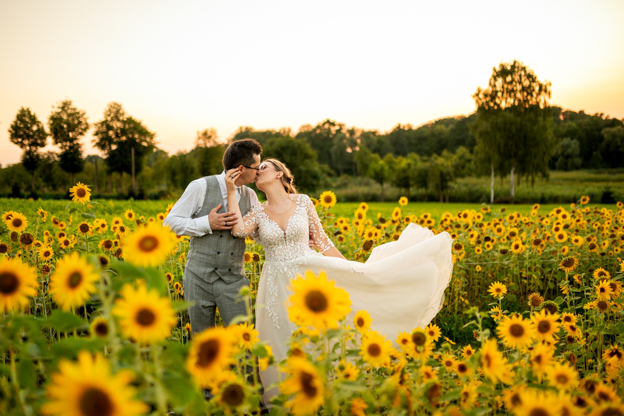 Pocałunek pary młodej w polu słoneczników o zachodzie słońca. Pan w szarej kamizelce, pani w białej sukni z koronkowymi rękawami. Romantyczna sesja plenerowa.