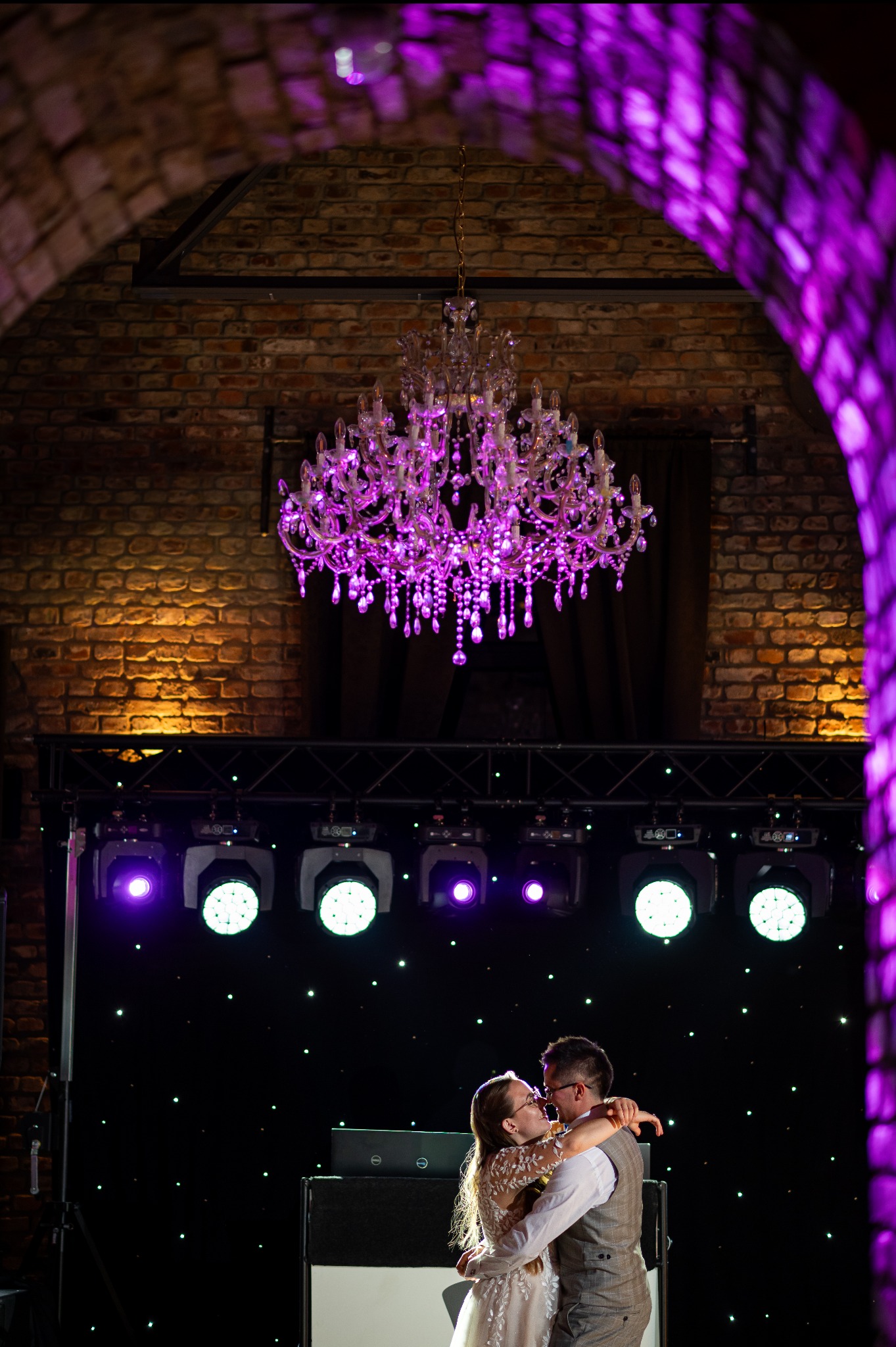 Para młoda tańczy w objęciach pod kryształowym żyrandolem, w tle ceglany łuk i oświetlenie sceniczne w odcieniach fioletu. Romantyczna atmosfera.