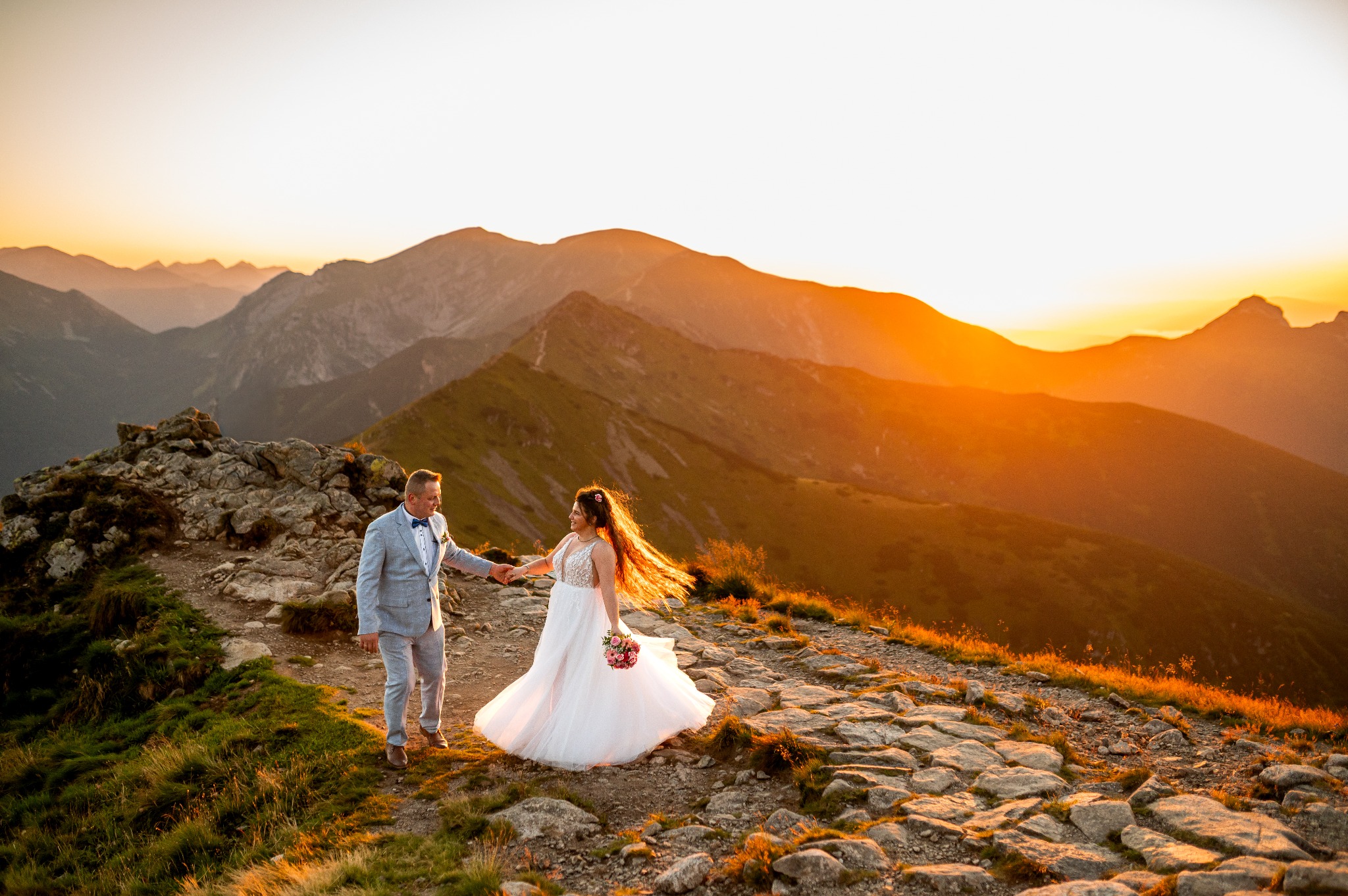 Para młoda w Zatorze na tle gór o zachodzie słońca. Panna młoda w białej sukni, pan młody w szarym garniturze. Kamienista ścieżka i ciepłe światło.