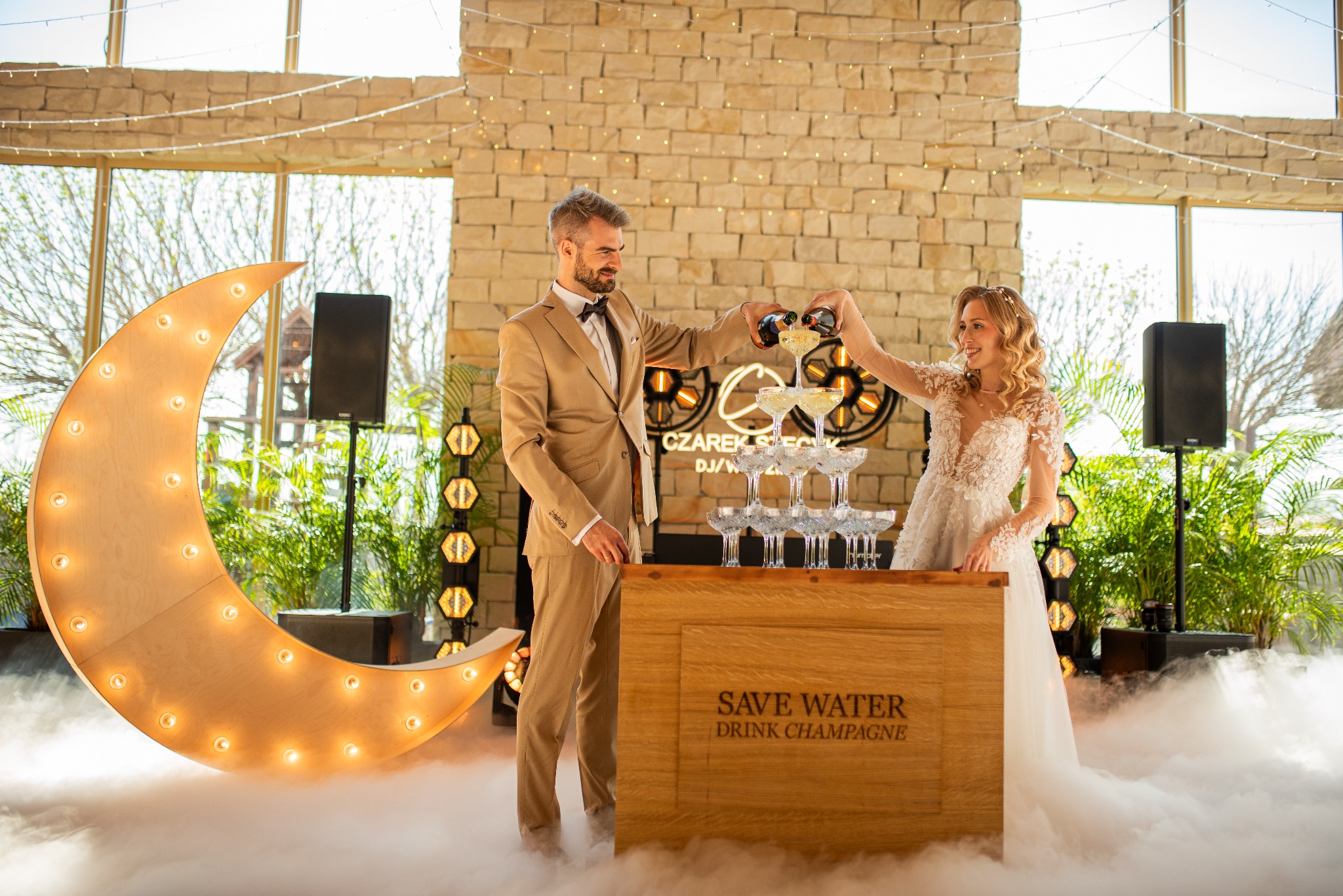 Para młoda w trakcie wznoszenia toastu szampanem, piramida kieliszków, dekoracje świetlne, napis 'Save Water Drink Champagne', efekt mgły.