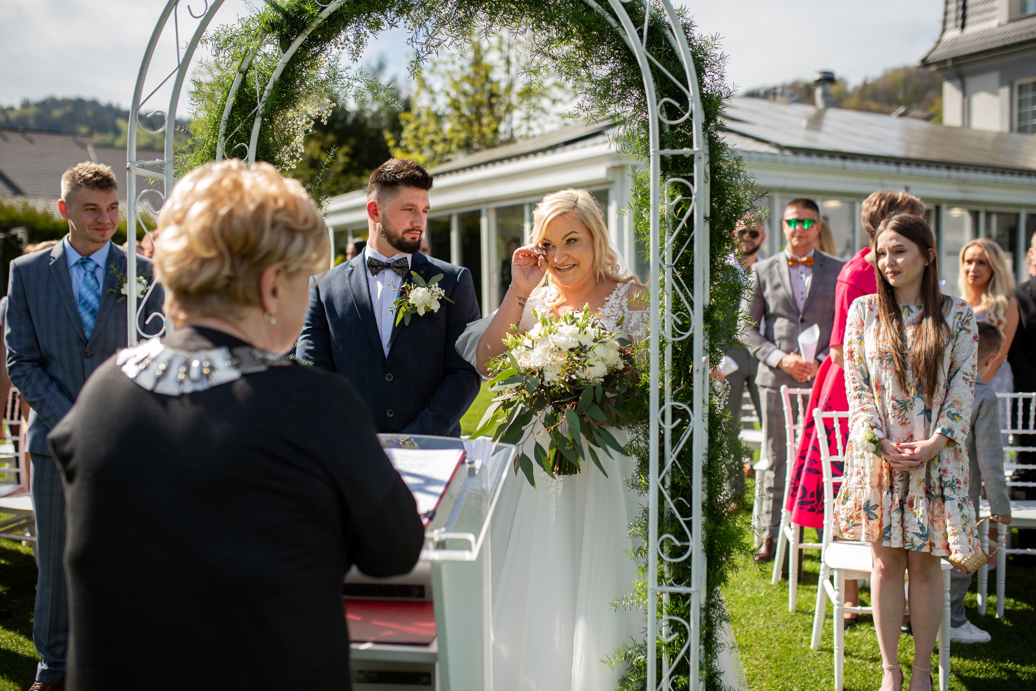 Wzruszona panna młoda z bukietem ślubnym podczas ceremonii plenerowej pod łukiem z zielenią, w tle goście i budynek z panelami słonecznymi.