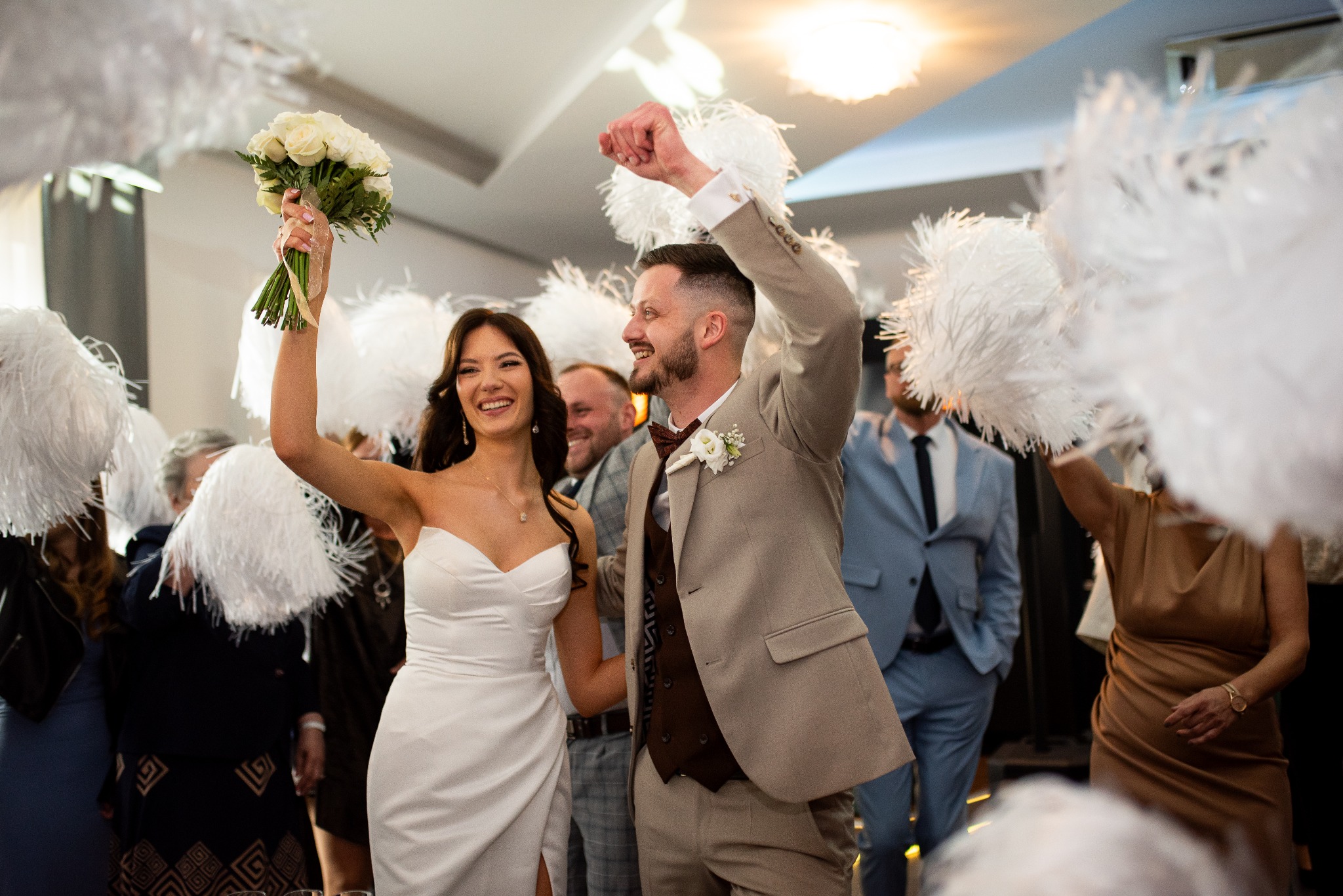 Rozradowana para młoda, on w beżowym garniturze i bordowej muszce, ona w białej sukni, wznoszą ręce z bukietem i pomponami na weselu, uśmiechnięci goście w tle.