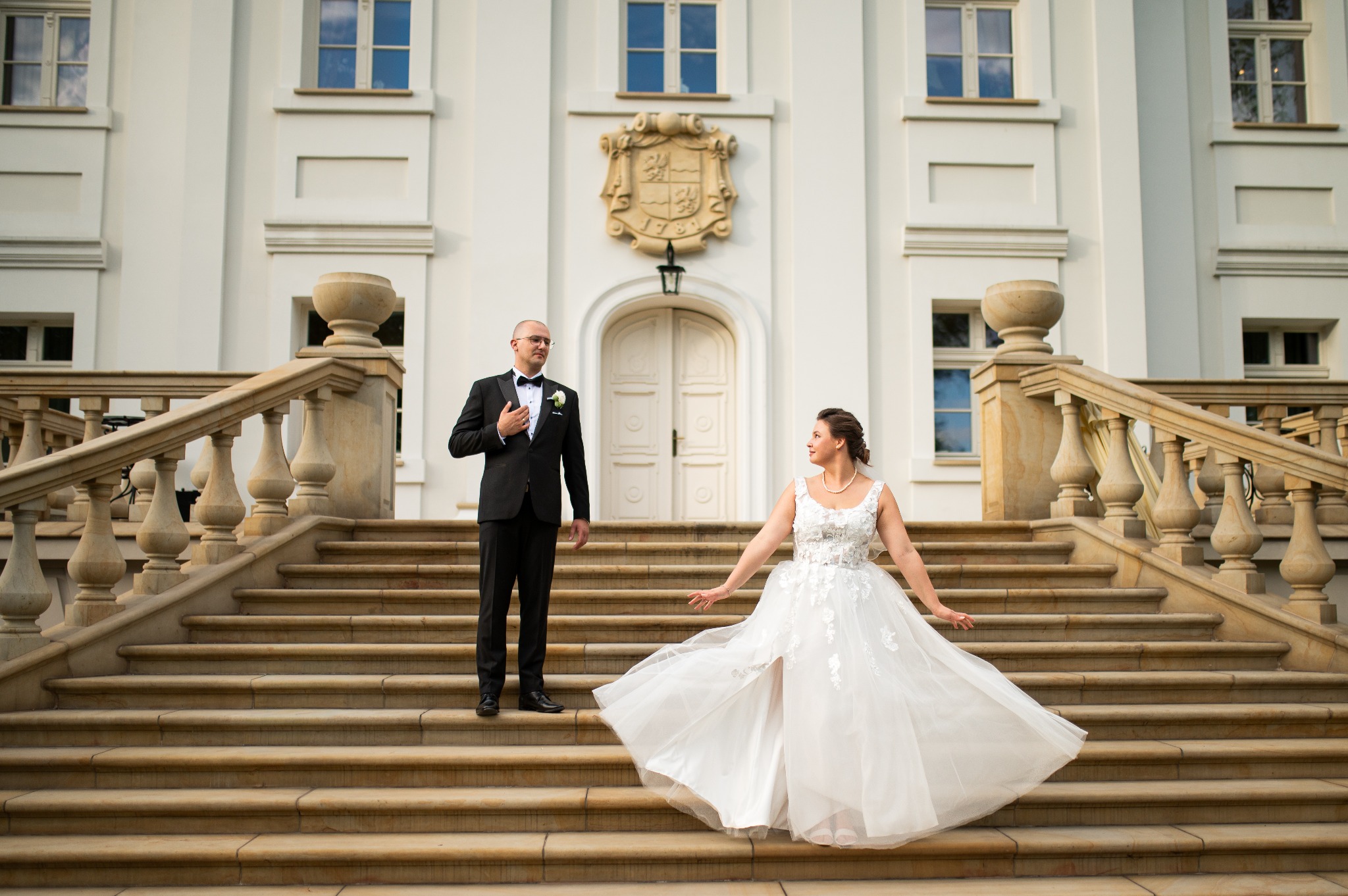 Para młoda na schodach pałacu z herbem, on w czarnym smokingu, ona w białej sukni z rozcięciem, w tle biała fasada budynku z oknami.