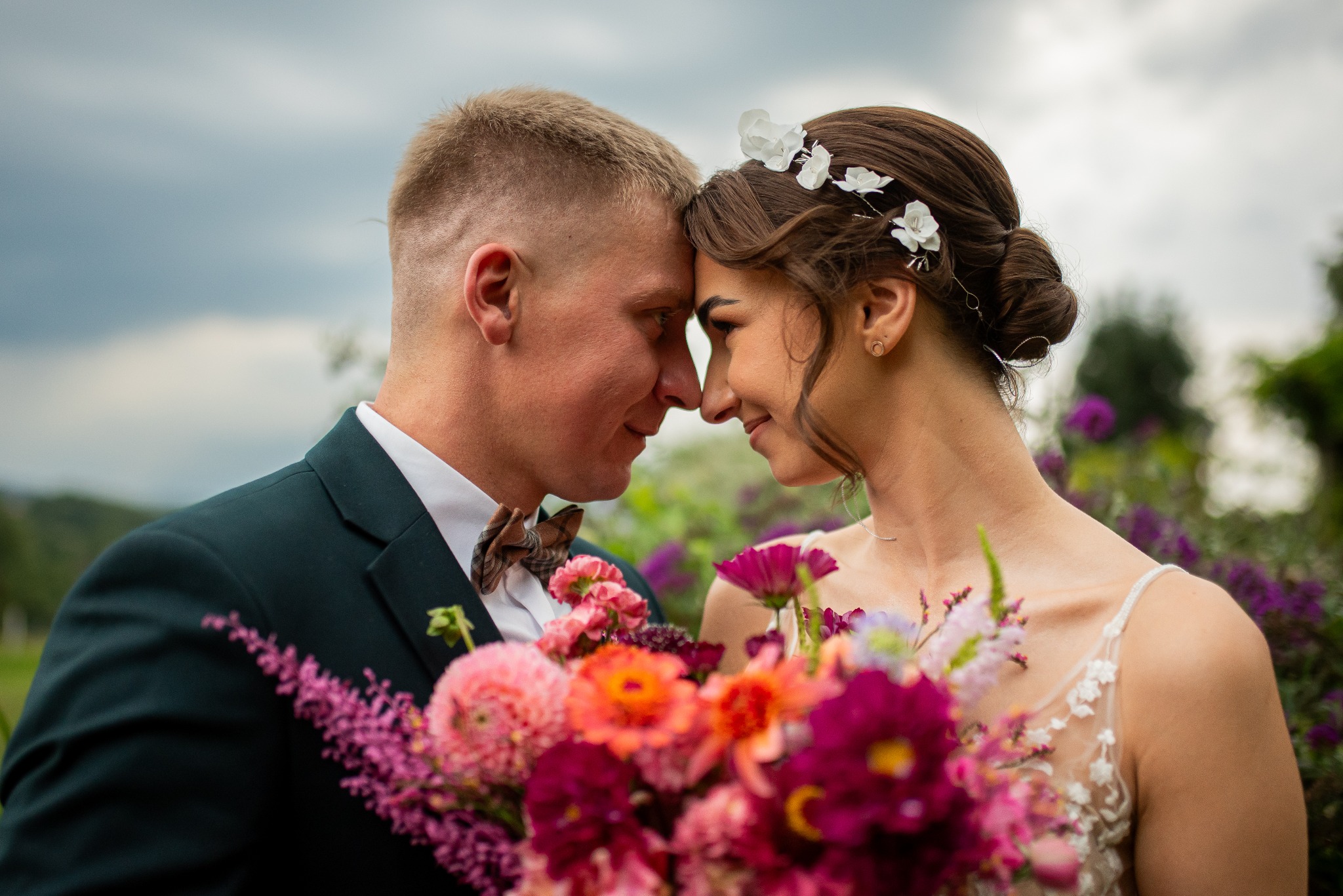 Para młoda w zbliżeniu, dotykająca się nosami. Ona w sukni z kwiatami, on w ciemnozielonym garniturze i muszce. Bukiet kwiatów na pierwszym planie.