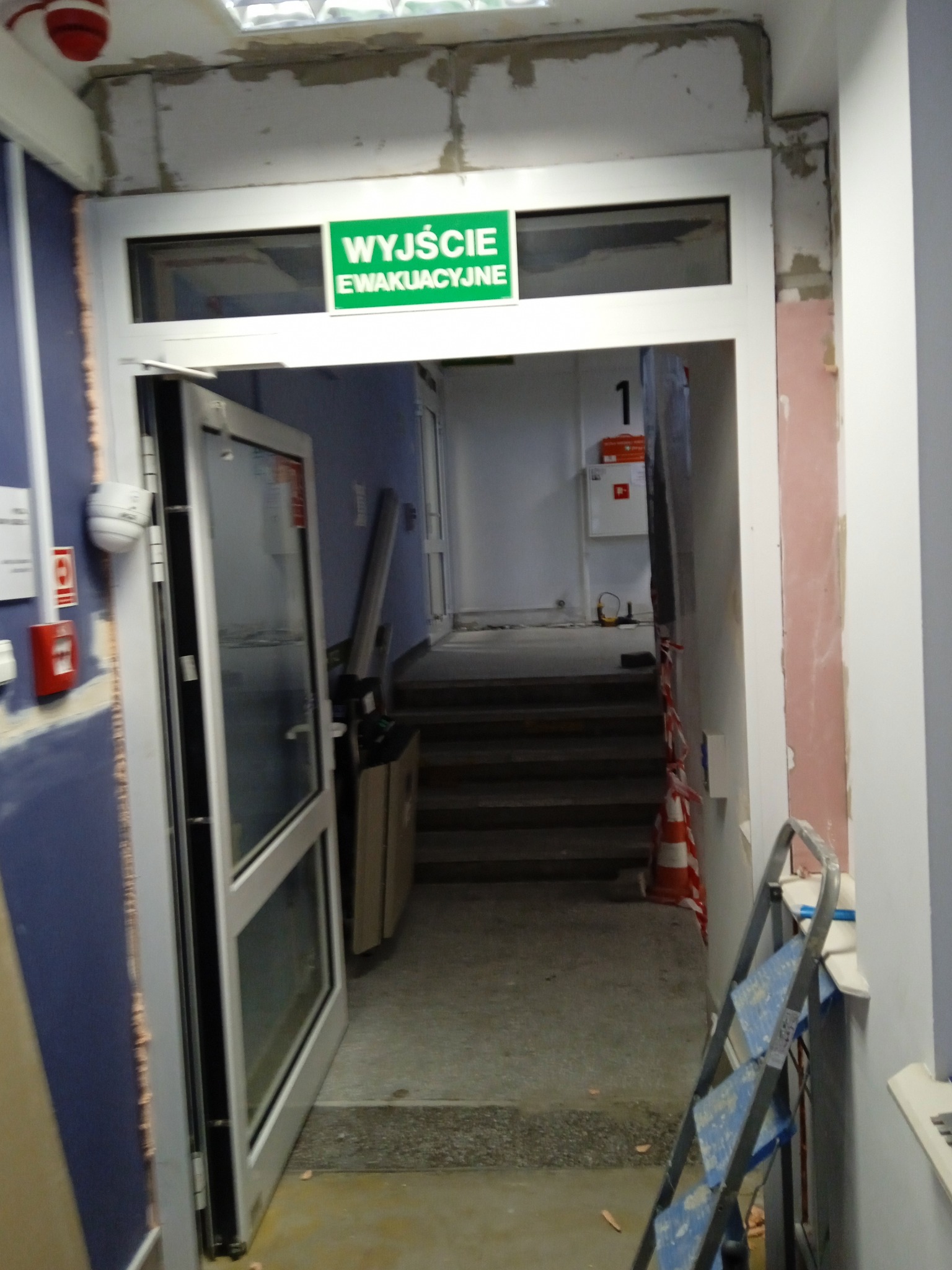 Korytarz w trakcie prac wykończeniowych: ściany częściowo pomalowane, widoczna instalacja nad drzwiami, drabina malarska, oznaczenie wyjścia ewakuacyjnego.