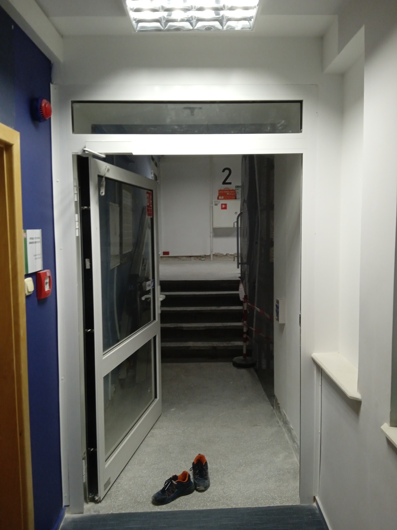 Otwarta aluminiowa rama drzwiowa z szybą nadświetlną, widok na schody i parę butów sportowych. Ściana pomalowana na niebiesko z elementami instalacji przeciwpożarowej.