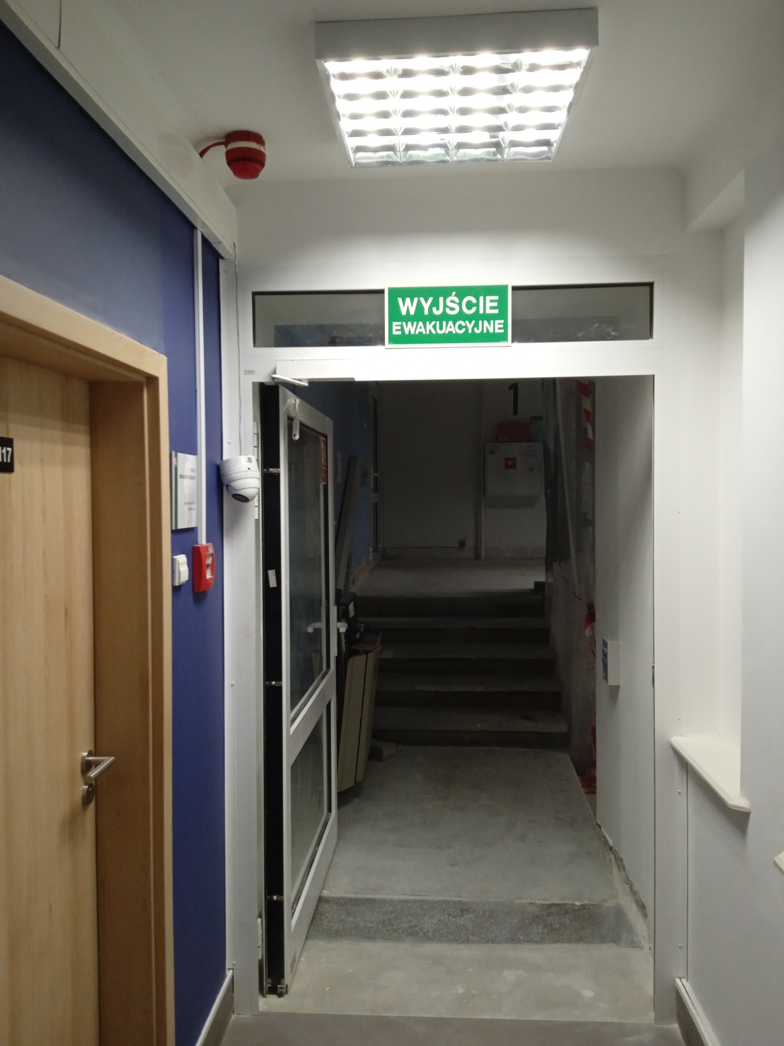 Korytarz ewakuacyjny z niebieską ścianą, drewnianymi drzwiami i zielonym znakiem 'Wyjście Ewakuacyjne' nad otwartymi drzwiami prowadzącymi na schody.
