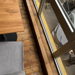 REMCOM EUROPE SPÓŁKA Z OGRANICZONĄ ODPOWIEDZIALNOŚCIĄ - Drewniana podłoga z widocznym fragmentem okna i stolika w lokalu usługowym, z zewnątrz widoczny zaparkowany samochód.