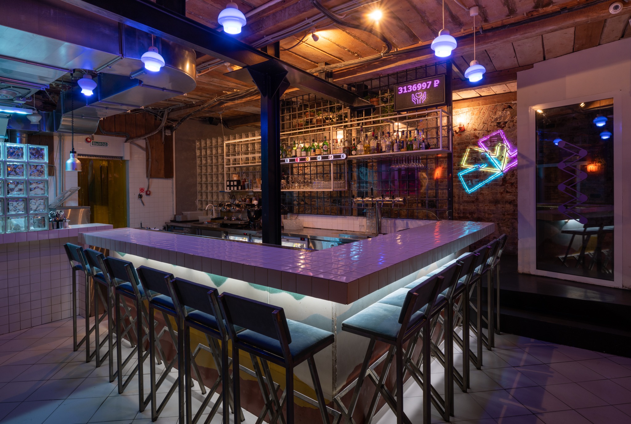 Nowoczesny bar z białym blatem z płytek, podświetlonym od spodu, oraz rzędem wysokich krzeseł z metalowymi nogami i niebieskimi siedziskami, oświetlony niebieskimi lampami i neonami.