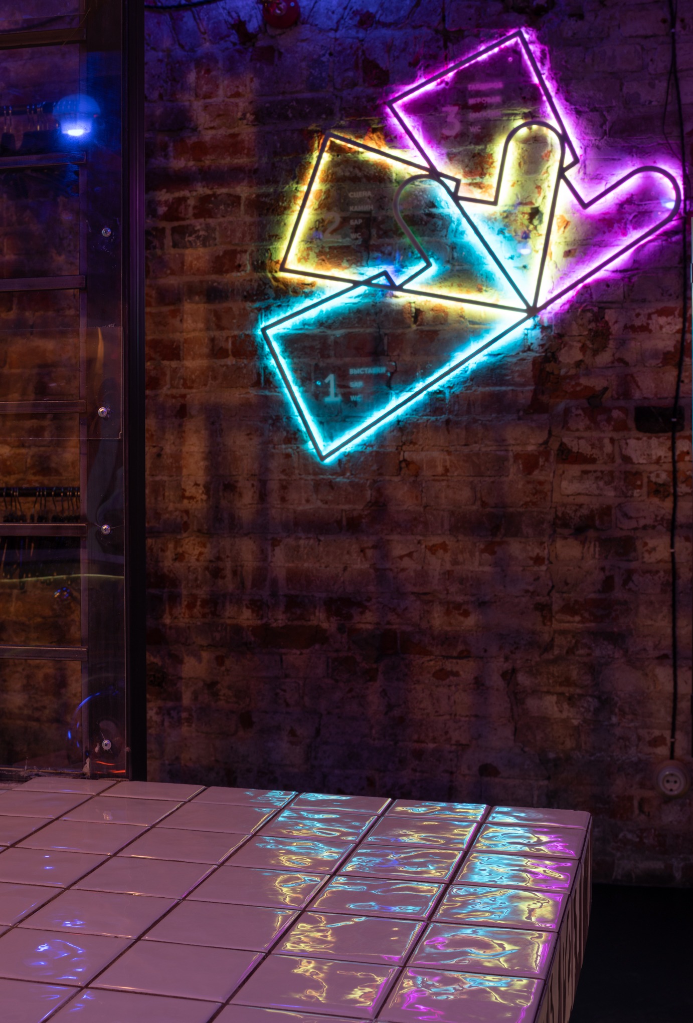 Neonowy znak z geometrycznymi kształtami na tle ceglanej ściany, odbijający się w błyszczących, białych płytkach posadzkowych.
