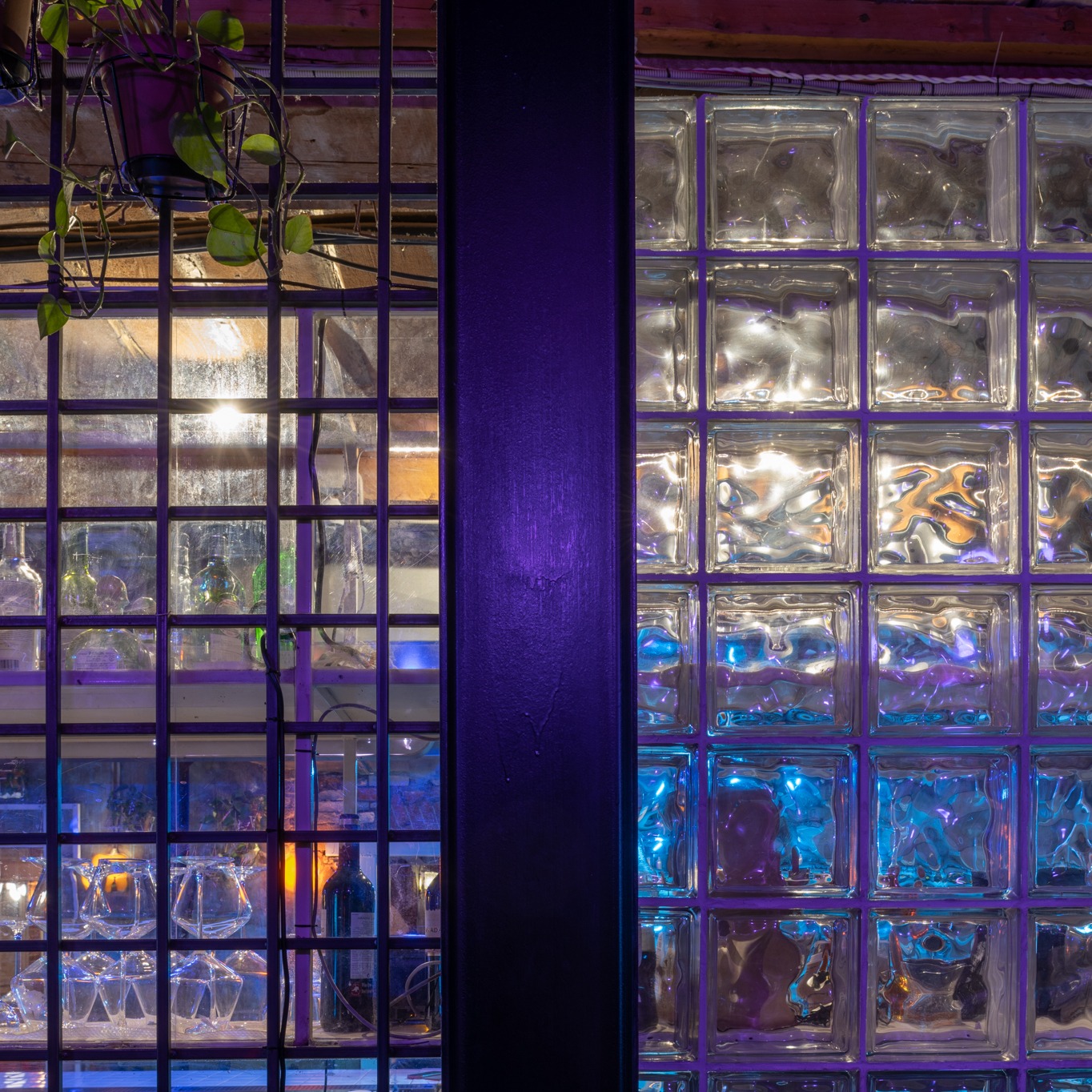 Wnętrze baru z ścianą z luksferów oświetloną niebieskim światłem i półką z kieliszkami oraz butelkami za metalową kratą, oświetloną ciepłym światłem.