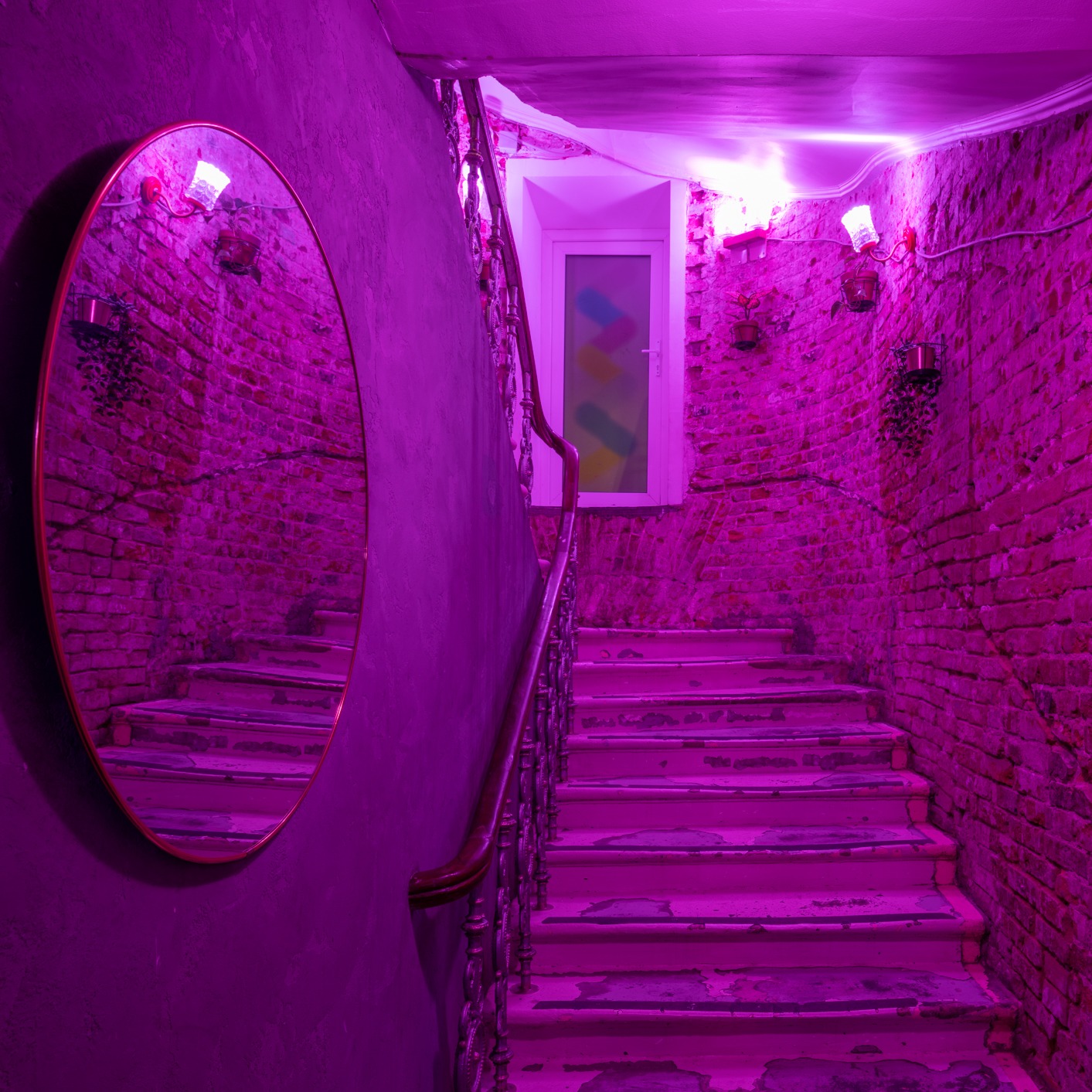 Klatka schodowa z ceglanymi ścianami i okrągłym lustrem, oświetlona intensywnym fioletowym światłem, widoczne schody z odpryskami farby i balustrada.