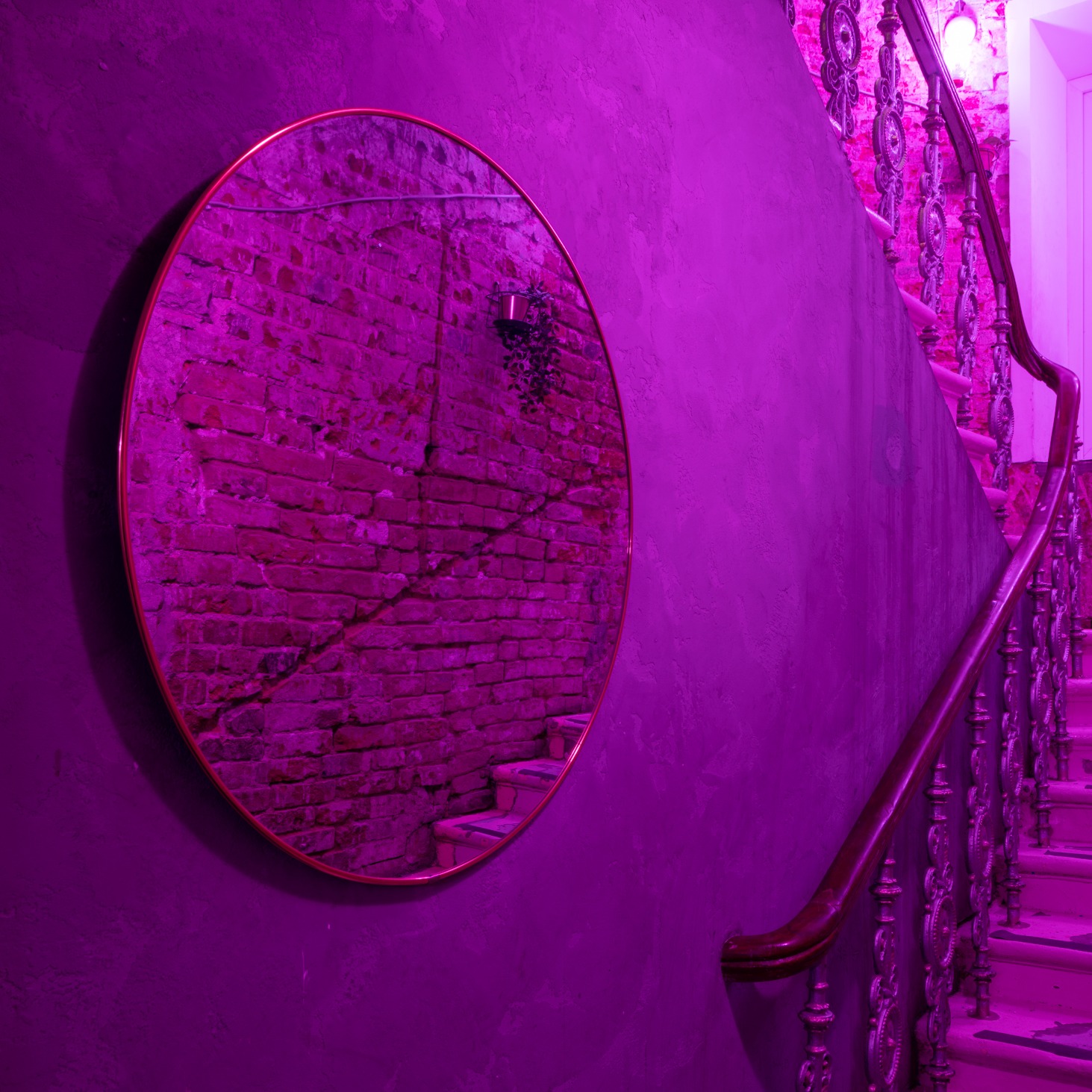 Okrągłe lustro z odbiciem ceglanej ściany i schodów, zawieszone na fioletowej ścianie w klatce schodowej w Warszawie, oświetlone fioletowym światłem.