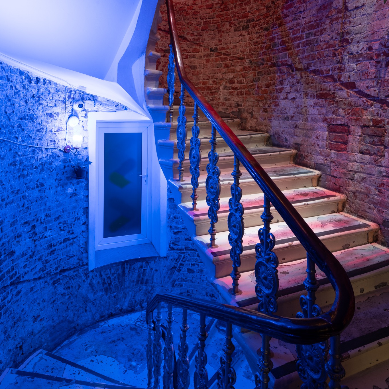 Spiralne schody z kutą balustradą, oświetlone niebieskim światłem, z fragmentem ceglanej ściany w tle.