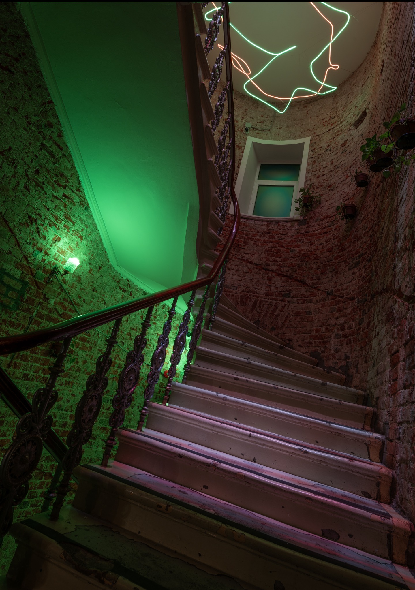 Spiralne schody z metalową balustradą w kamienicy z ceglanymi ścianami, oświetlone zielonym światłem, z neonowym oświetleniem sufitowym i roślinami doniczkowymi na ścianach.