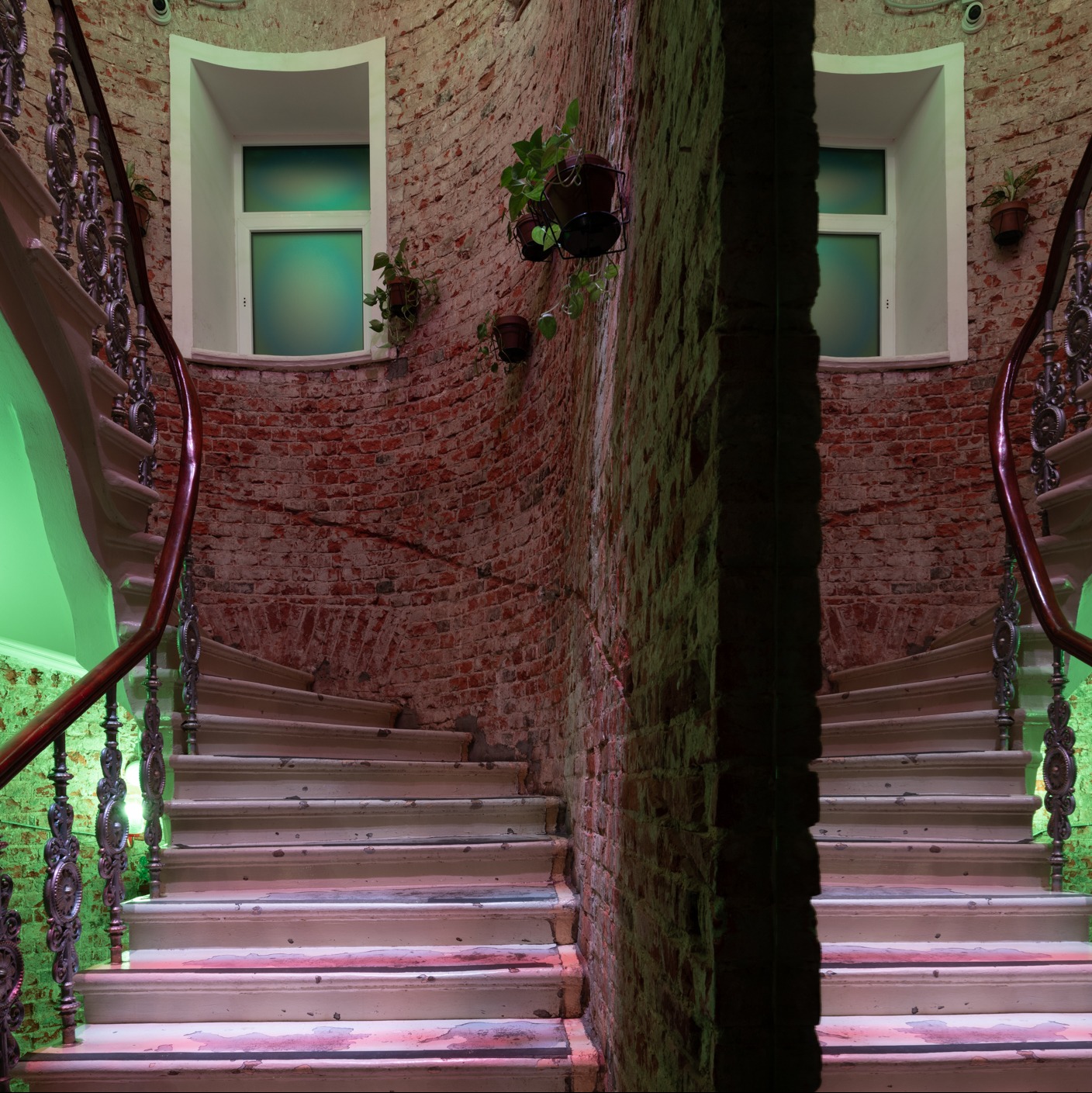 Spiralne schody z kutą balustradą w klatce schodowej z ceglanymi ścianami i oknami z zielonkawym szkłem, ozdobione wiszącymi roślinami w doniczkach.
