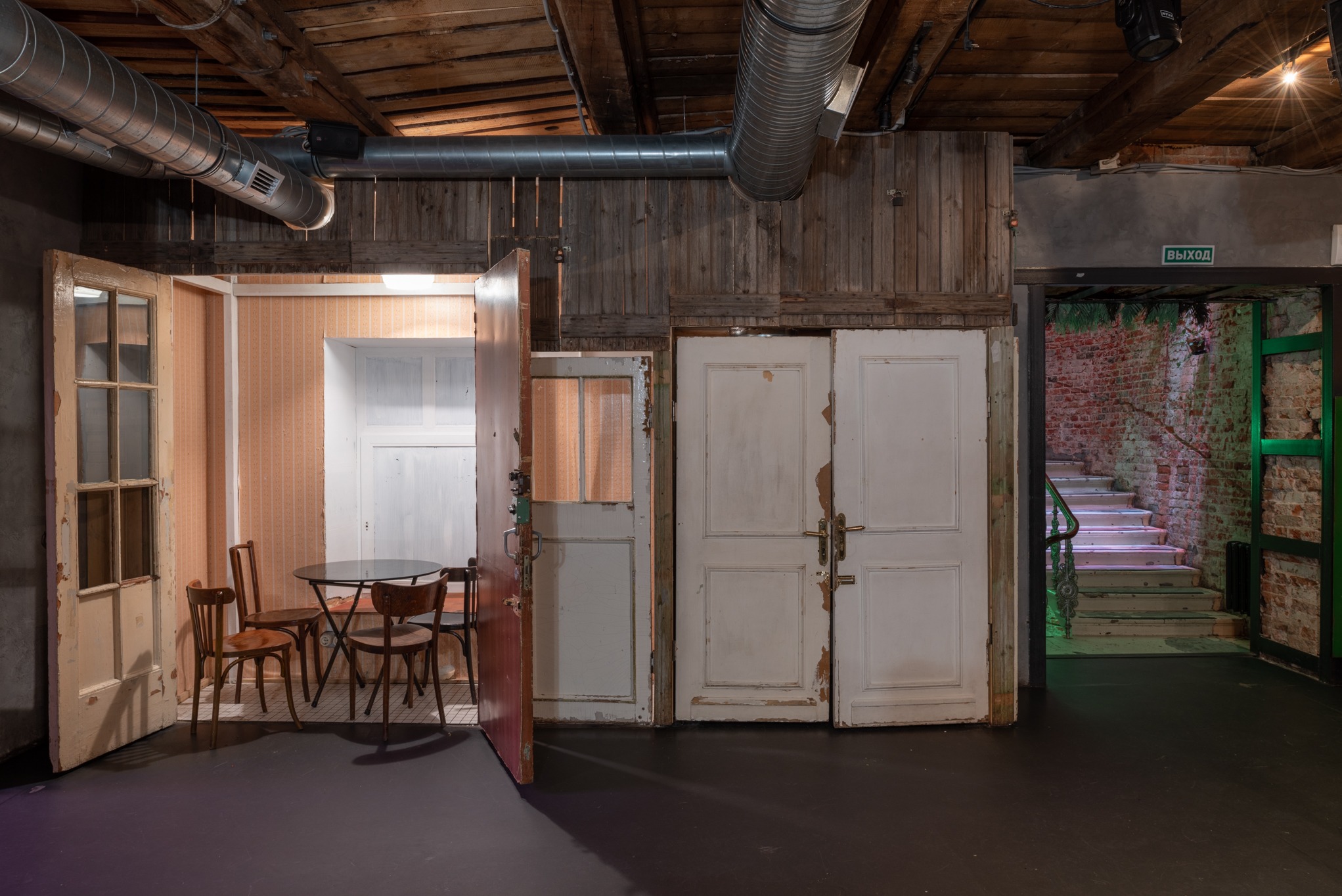 Wnętrze z drewnianym stropem, odsłoniętymi rurami wentylacyjnymi i różnymi drzwiami, z których jedne prowadzą do małego pokoju z tapetą i stolikiem z krzesłami, a inne na klatkę schodową...