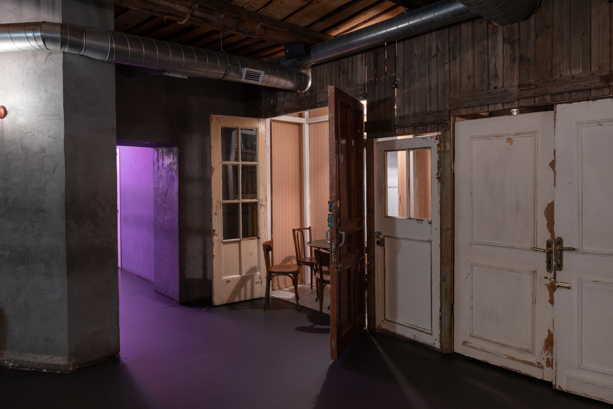 Wnętrze pomieszczenia w trakcie prac wykończeniowych; widoczne surowe ściany, odsłonięte drewniane belki stropowe, instalacja wentylacyjna oraz stare, częściowo odrestaurowane drzwi z widocznymi...