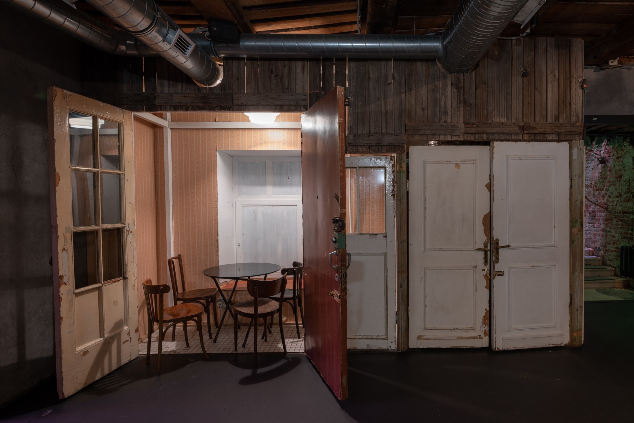 Ustawiona scena z wykorzystaniem starych drzwi i mebli: pokój z tapetą w paski, okrągły stolik z krzesłami, widoczne rury wentylacyjne pod drewnianym stropem.