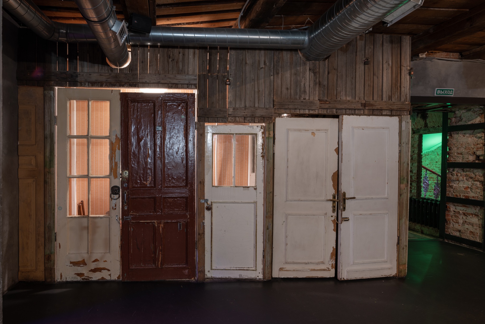 Różne, odrestaurowane i nieodrestaurowane stare drzwi drewniane w pomieszczeniu z drewnianymi ścianami i widocznymi rurami wentylacyjnymi pod sufitem. Jedne drzwi są brązowe, inne białe,...