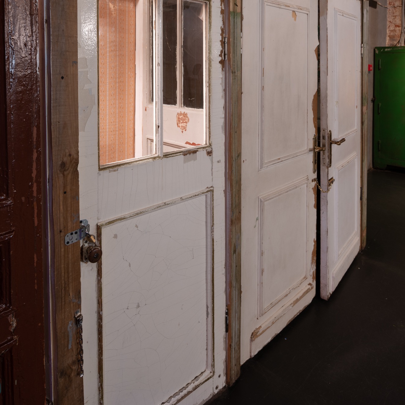 Stare, białe drzwi z widocznymi śladami zużycia, odpryskującą farbą i elementami drewnianymi, częściowo otwarte, ukazujące fragment wnętrza z wzorzystą tapetą, zamek i klamka w stylu retro.