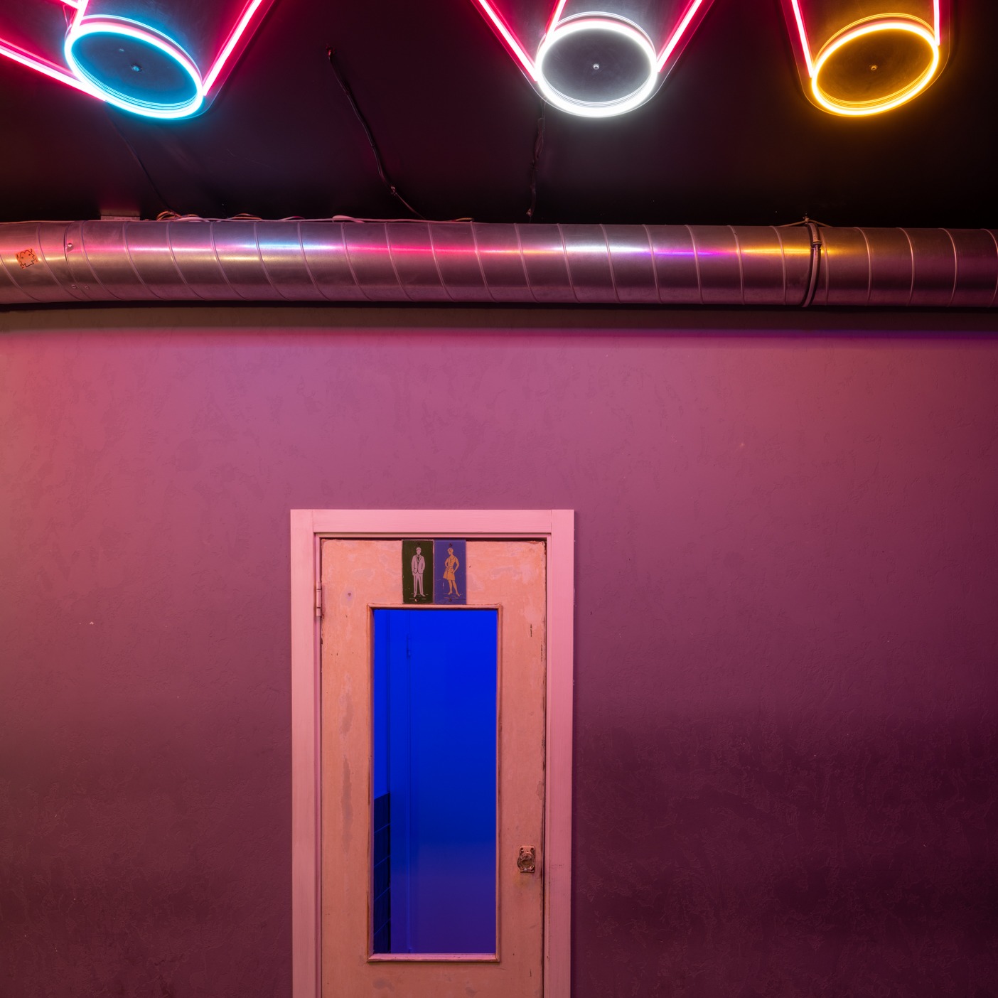 Pomieszczenie z fioletową ścianą, rurą wentylacyjną pod sufitem z neonami i drzwiami z symbolem toalety damskiej i męskiej.