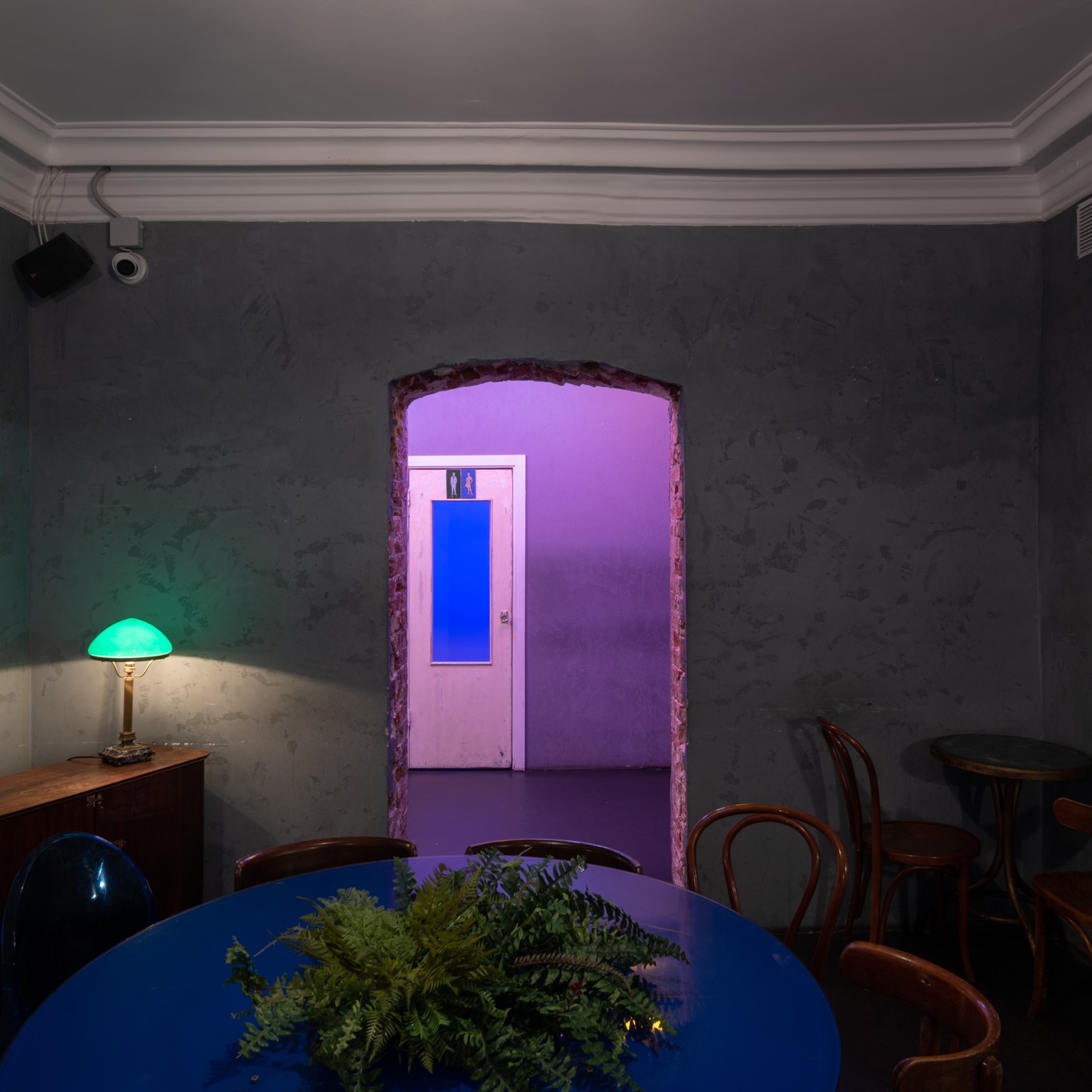 Wnętrze lokalu z surowymi szarymi ścianami, niebieskim okrągłym stołem z paprotką, zieloną lampką na komodzie i wejściem do toalety oświetlonym na fioletowo.
