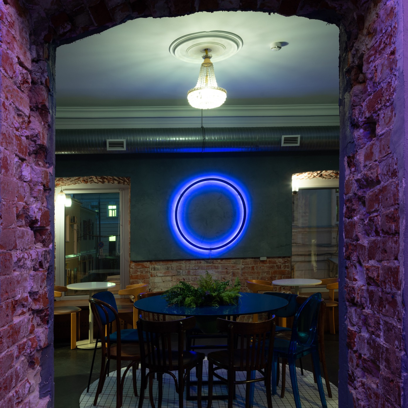 Wnętrze lokalu z ceglaną ścianą, okrągłym neonem, żyrandolem i stolikami z krzesłami, widoczne przez ceglane przejście.