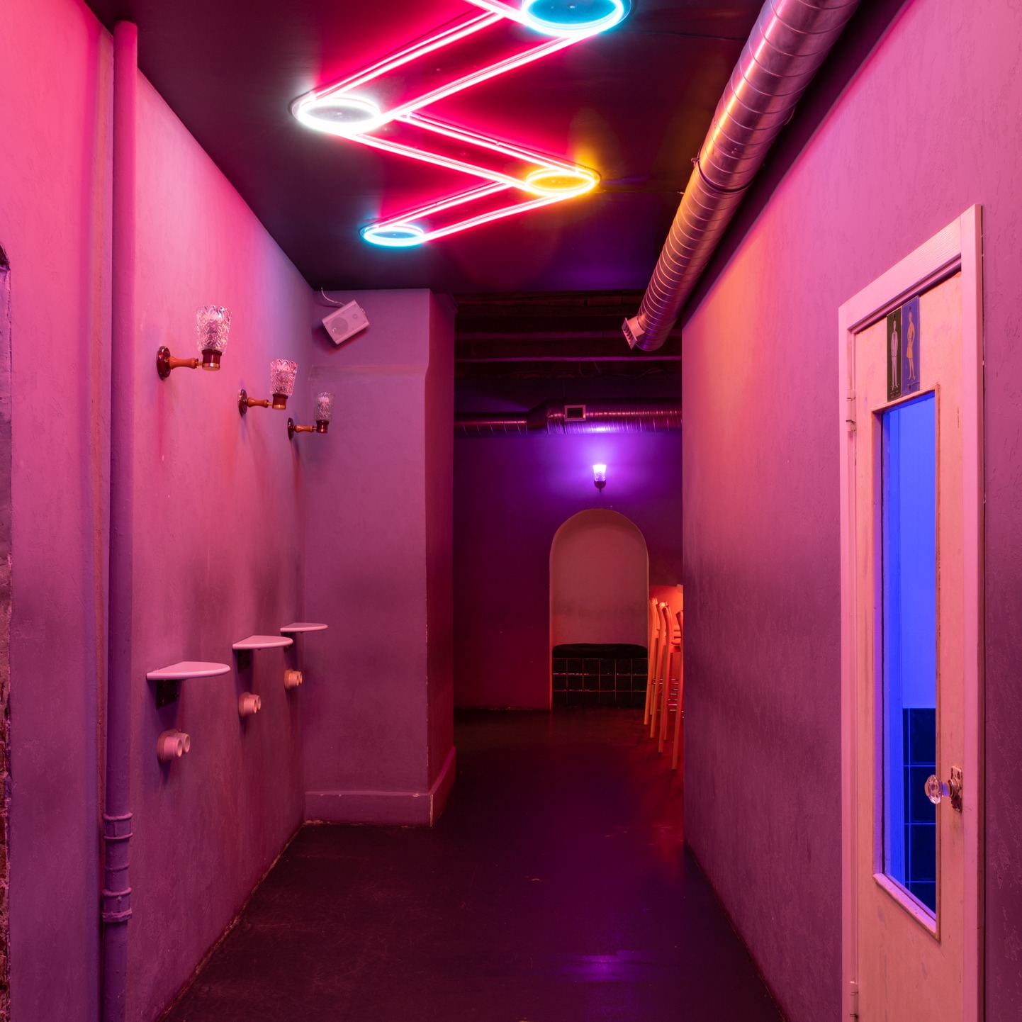 Korytarz oświetlony neonami w kształcie zygzaka i lampami ściennymi z kryształowymi kloszami, z trzema małymi półkami zamontowanymi na rurach, prowadzący do pomieszczenia z łukowatym wejściem...