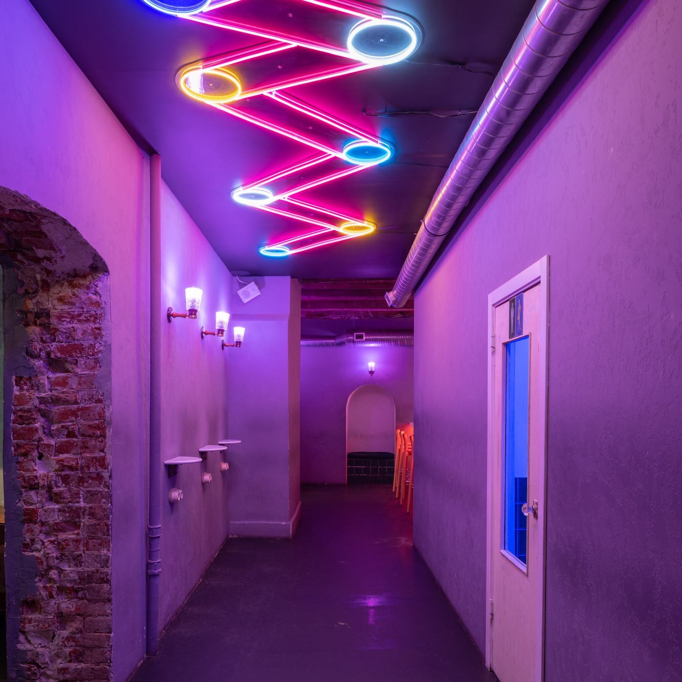 Długi korytarz oświetlony neonowymi lampami w kształcie zygzaka z okrągłymi elementami w kolorach różowym, niebieskim i żółtym, ściany w odcieniach fioletu, widoczna instalacja wentylacyjna...