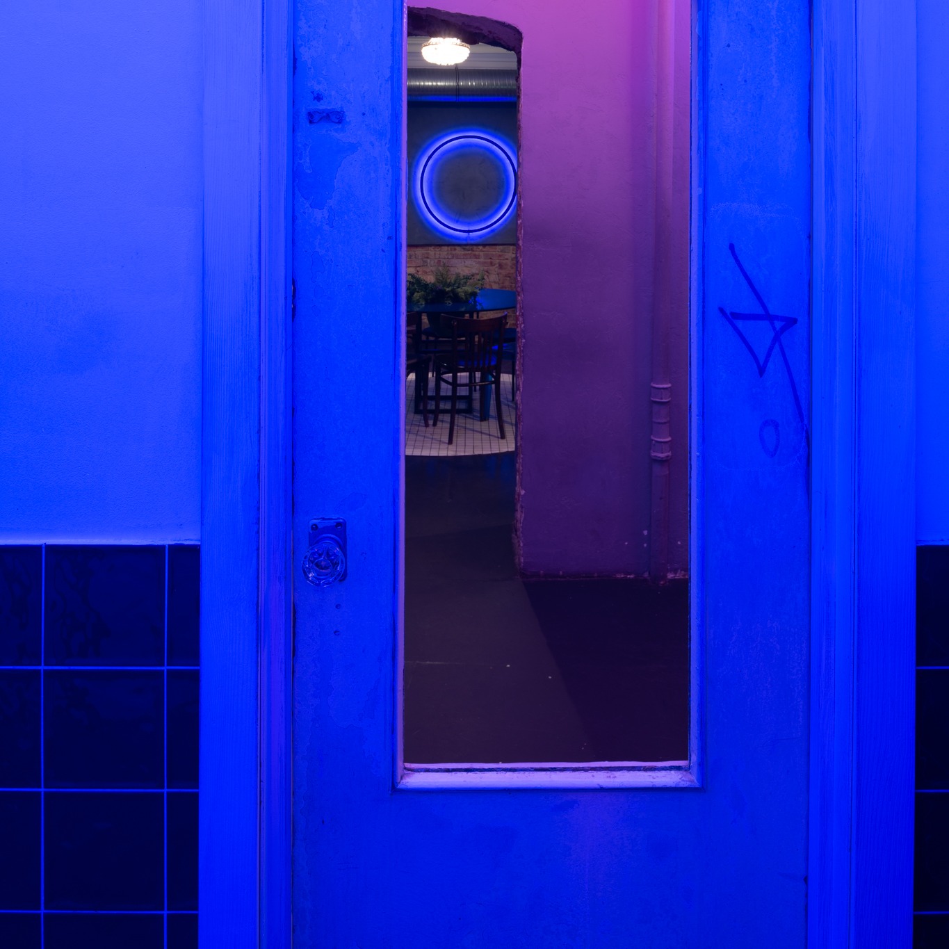 Oświetlona na niebiesko odnowiona łazienka z czarnymi kaflami i drzwiami z szybką, widok na dalszą część pomieszczenia z neonowym okręgiem na ścianie.