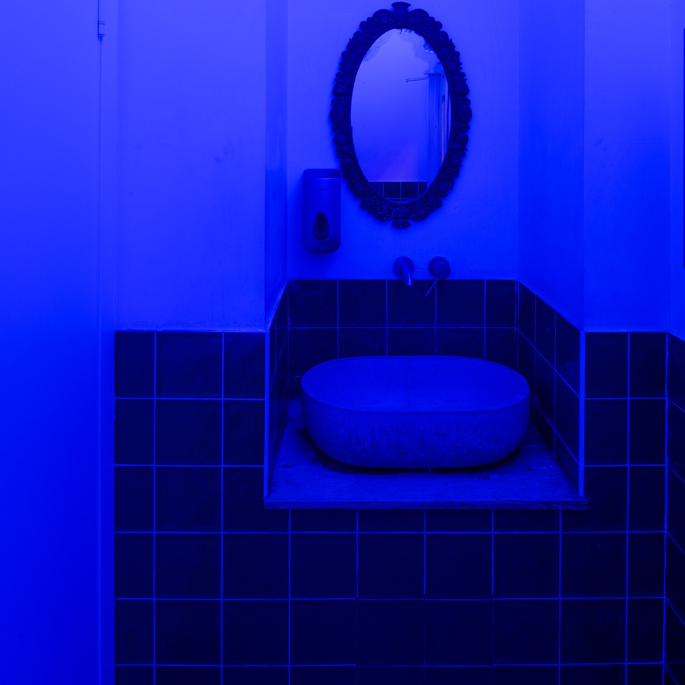 Niebieskie oświetlenie w łazience: umywalka nablatowa na drewnianej półce, ciemna glazura, ozdobne lustro i dozownik mydła.