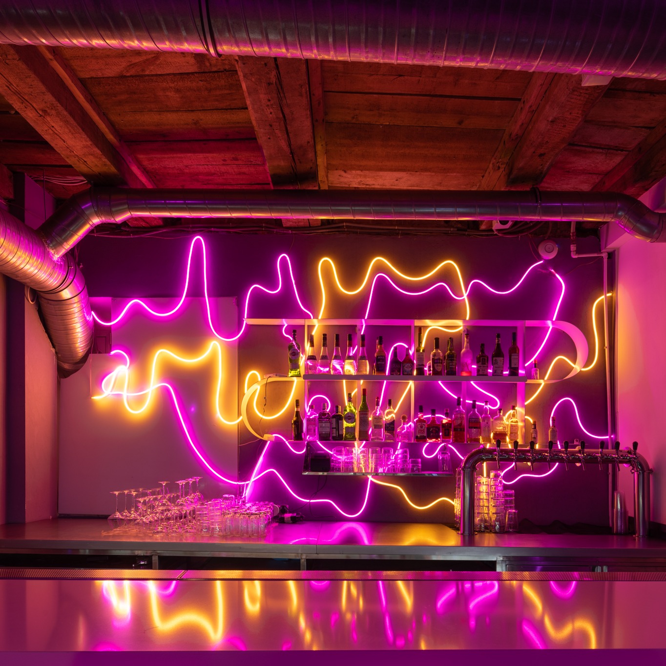 Nowoczesny bar z neonowym oświetleniem w odcieniach różu i żółci, butelki alkoholi na półkach, metalowy blat baru, widoczne rury wentylacyjne pod drewnianym sufitem.