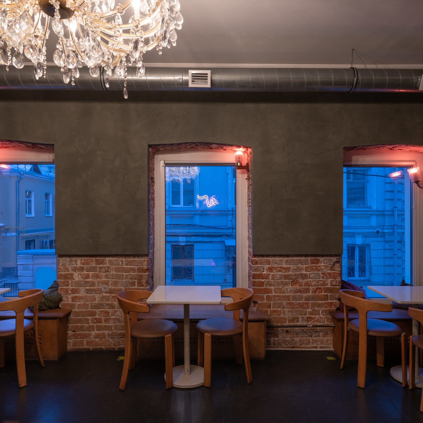 Wnętrze kawiarni z surowymi ścianami, ceglanymi elementami, drewnianymi krzesłami i stolikami, oświetlone kryształowym żyrandolem i neonami za oknami.