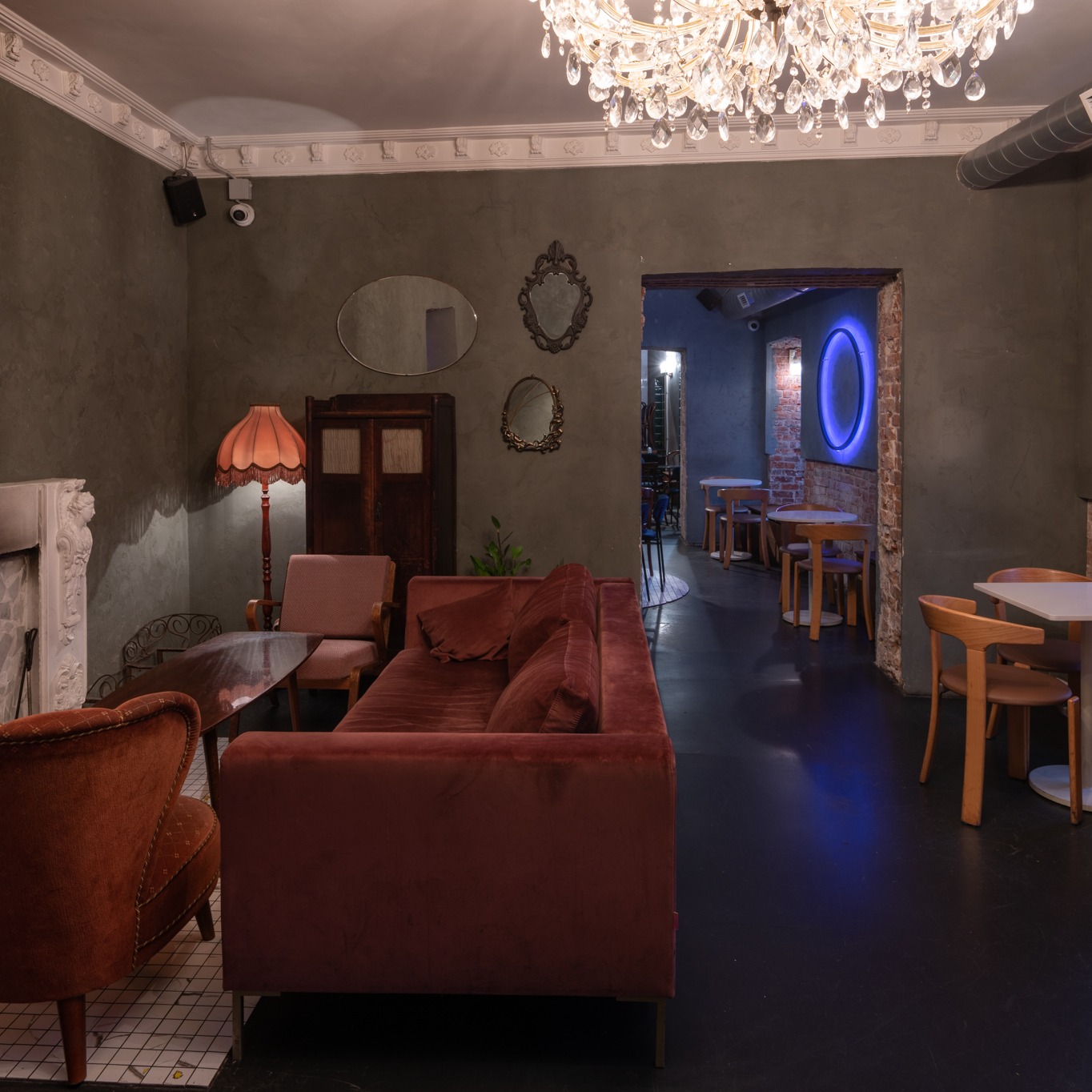 Wnętrze lokalu z elementami stylu retro: welurowa sofa, lampa z abażurem, lustra w ozdobnych ramach, kryształowy żyrandol i dekoracyjna sztukateria sufitowa.