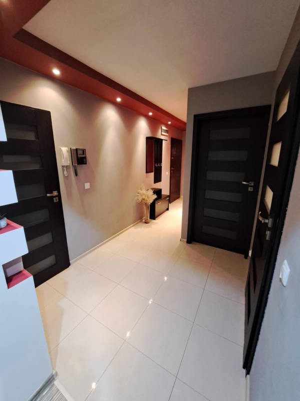 Jasny korytarz po remoncie z białymi płytkami na podłodze, ciemnymi drzwiami z poziomymi przeszkleniami i szarą ścianą z dekoracyjnym pasem w kolorze bordo pod sufitem.