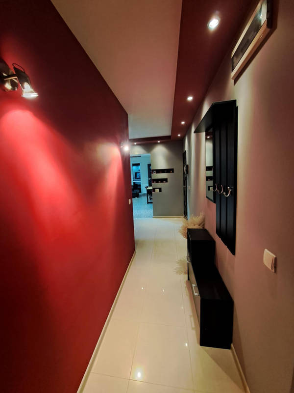 Pomalowany na czerwono korytarz z błyszczącą podłogą, oświetlony lampami ściennymi i punktowymi, z czarnymi meblami wzdłuż ściany i widokiem na dalsze pomieszczenia.