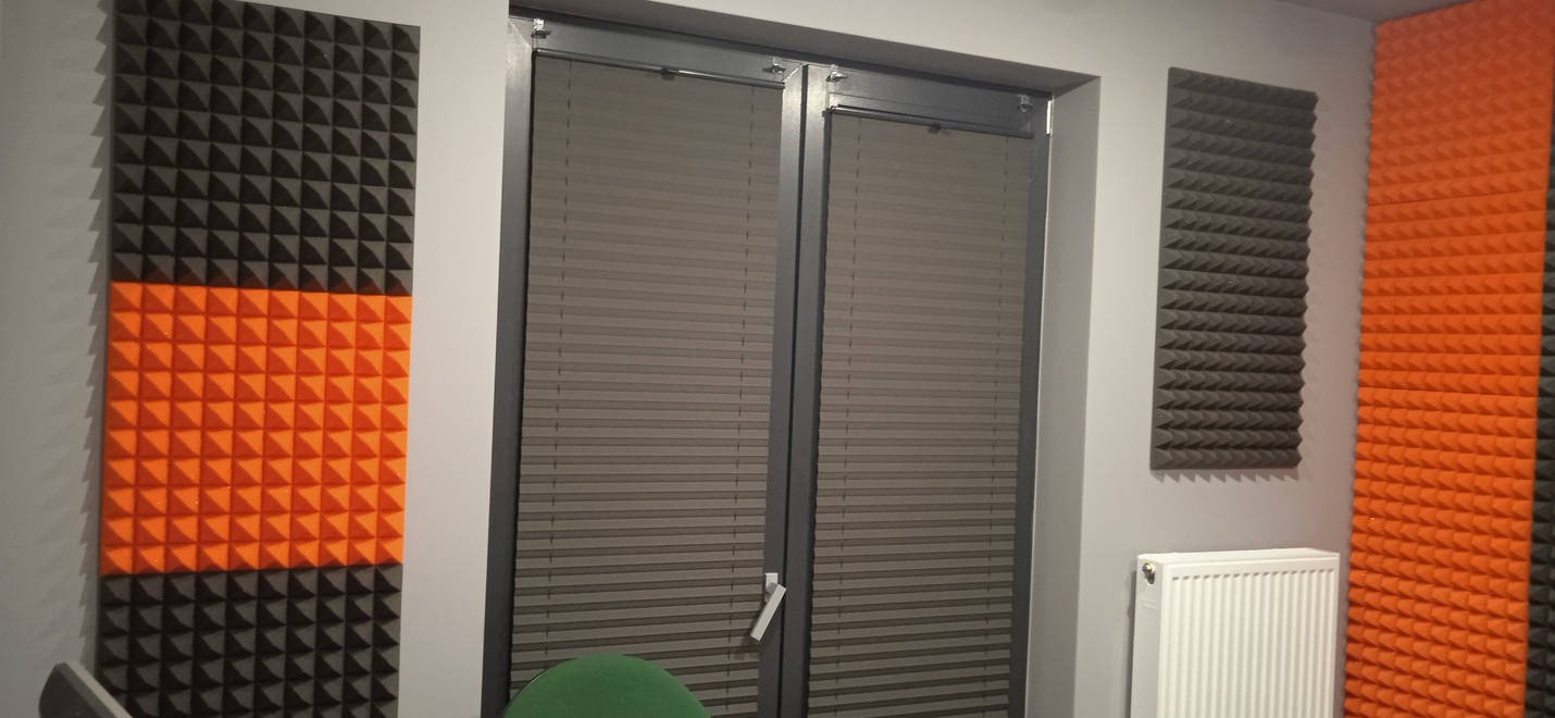 Pokój z panelami akustycznymi w kolorach czarnym i pomarańczowym, zamontowanymi na ścianach obok okna z żaluzjami i grzejnika.