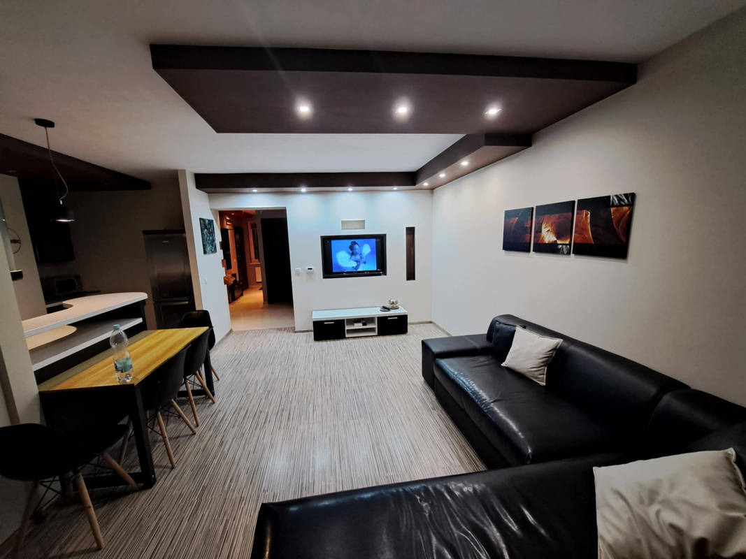 Salon po remoncie z czarną skórzaną sofą, telewizorem na ścianie i podwieszanym sufitem z oświetleniem punktowym, widok na otwartą kuchnię.