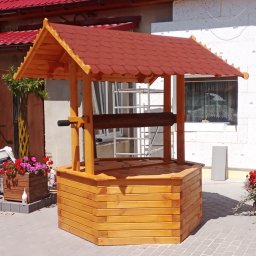 Jacek Czil - Drewniana studnia ogrodowa z czerwoną dachówką, widoczna z bliska na tle domu z oknem i rusztowaniem, obok donica z kwiatami.