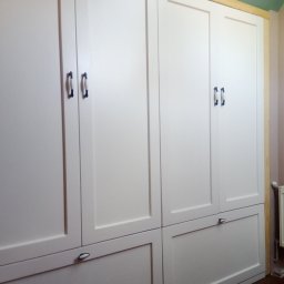 Jacek Czil - Biała, zabudowana szafa z drewnianymi frontami, ciemnymi uchwytami i widocznym fragmentem drewnianej ramy oraz podłogi.