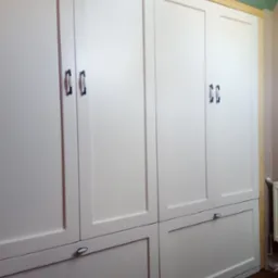 Biała, zabudowana szafa z drewnianymi frontami, ciemnymi uchwytami i widocznym fragmentem drewnianej ramy oraz podłogi.