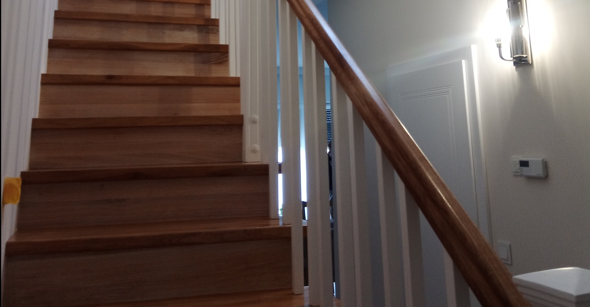 Fragment widoku na dębowe stopnie schodów z białymi balustradami, oświetlone kinkietem na ścianie, z widocznym fragmentem poręczy i drzwi w tle.