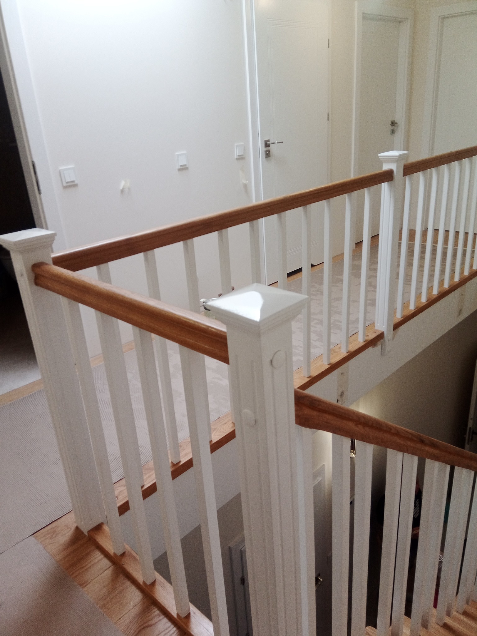 Nowoczesne schody wewnętrzne z białymi balustradami i drewnianym poręczą, widok z góry na korytarz z białymi drzwiami i jasnym oświetleniem.