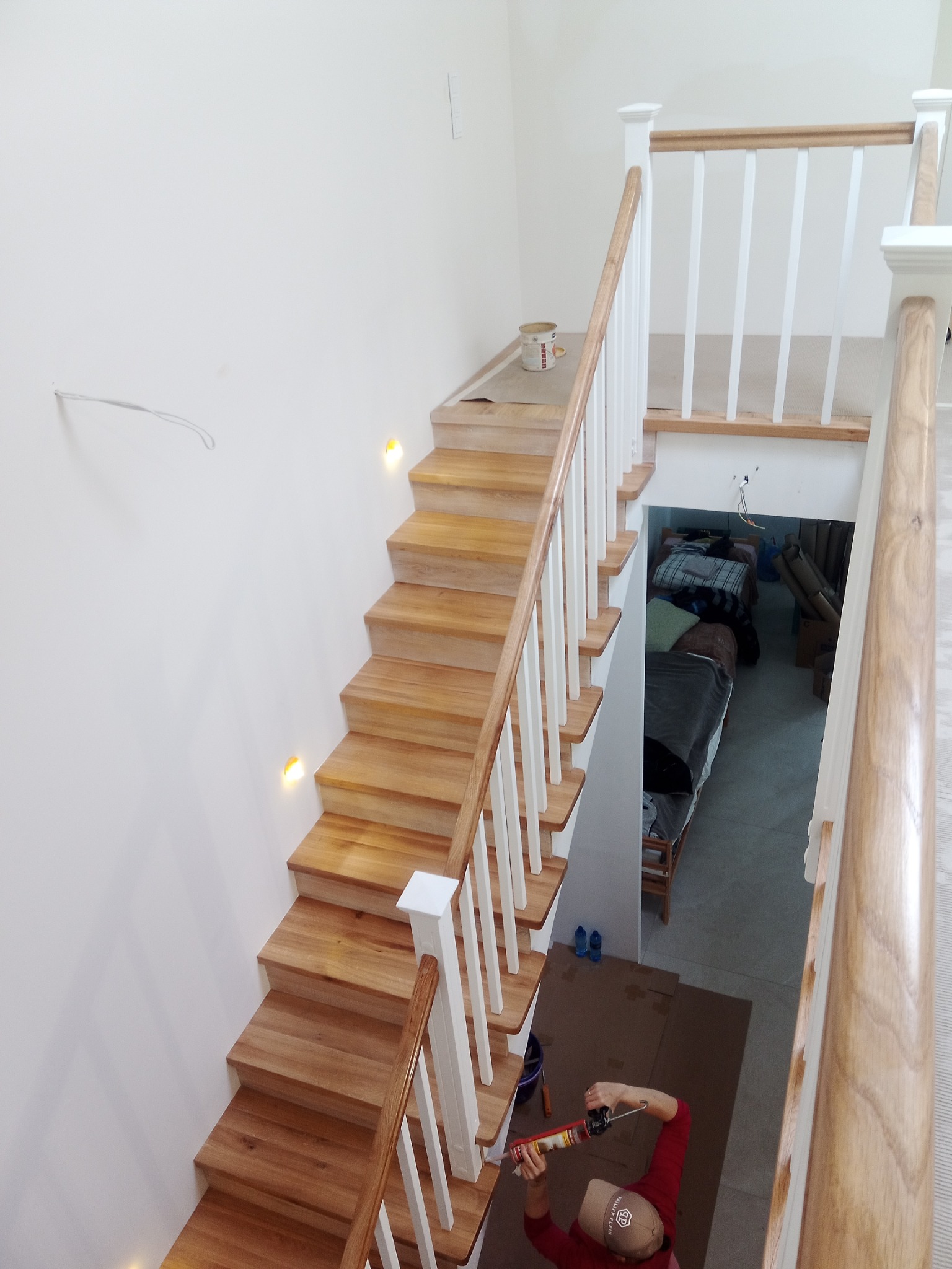 Montaż drewnianych schodów z białymi balustradami, widok z góry na pracownika aplikującego uszczelniacz, w tle zagospodarowana przestrzeń pod schodami.