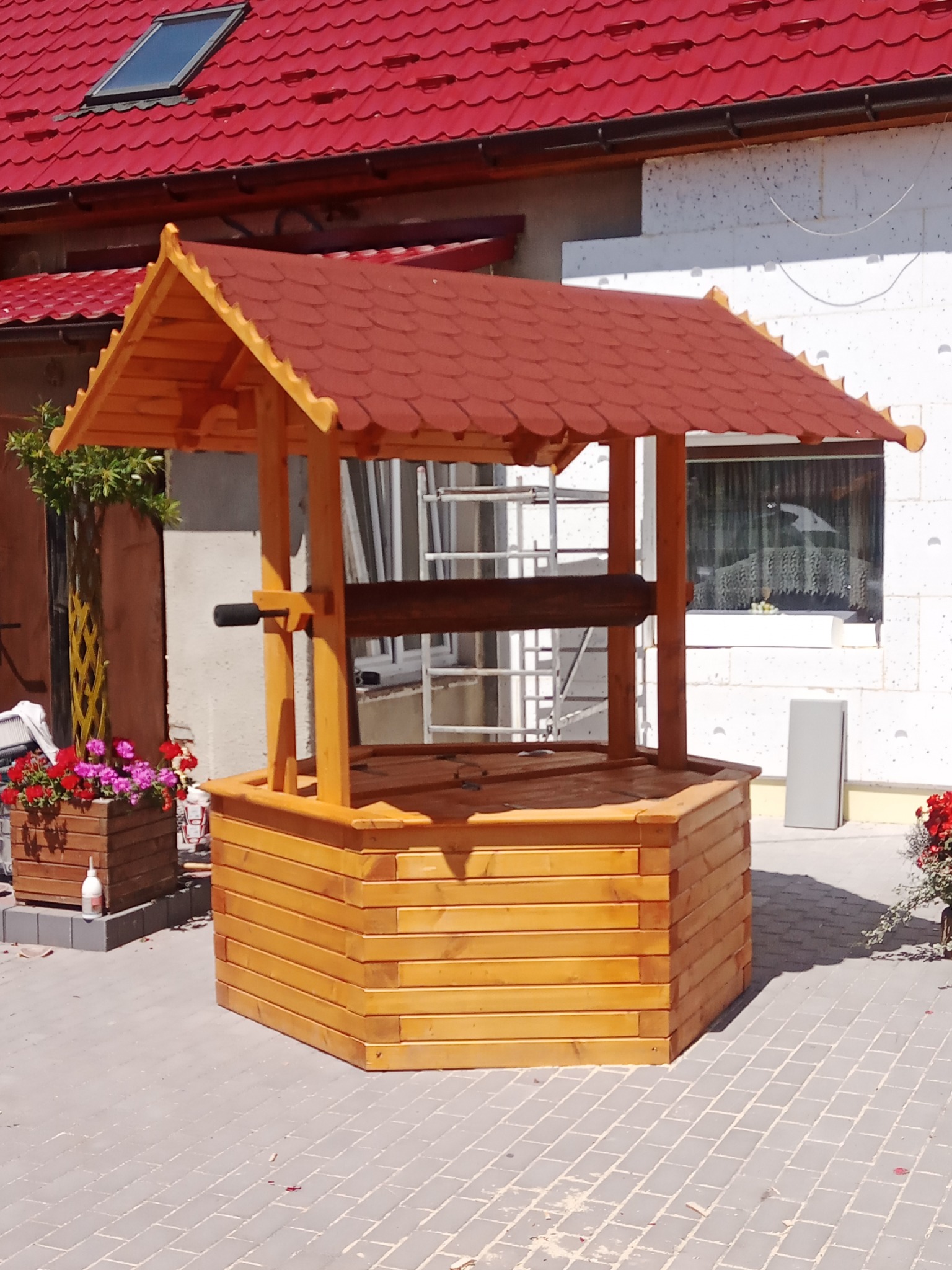 Drewniana studnia ogrodowa z czerwoną dachówką, widoczna z bliska na tle domu z oknem i rusztowaniem, obok donica z kwiatami.